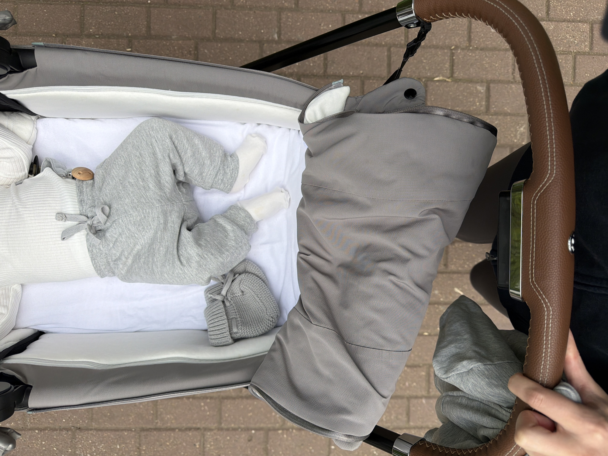 A top-down view captures a baby lying in a stroller. The baby is dressed in a white top and gray pants, along with white socks. Next to the baby's legs is a gray knitted hat. The stroller is equipped with a gray footmuff. A person's hands are visible, gripping the brown leather-covered handle of the stroller. The stroller is on a brick sidewalk. The location is inferred, but not definitively identifiable from the image, as Khimki, Russia.