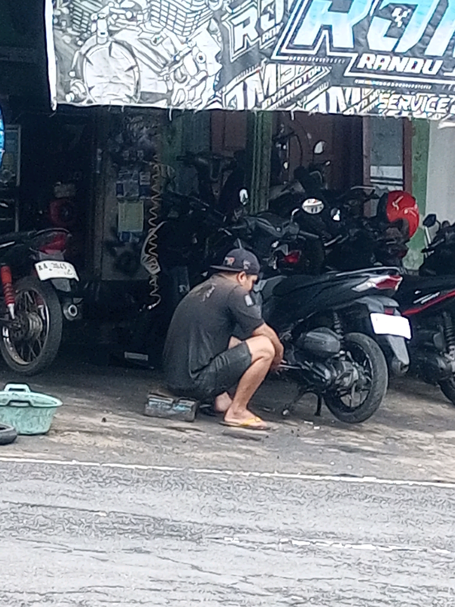 The image shows a person working on a motorcycle in what appears to be an outdoor workshop or garage in Keseneng, Indonesia. The person, wearing a dark t-shirt, dark shorts, and a baseball cap, is kneeling beside a black motorcycle, with their hands near the engine. A red helmet is visible on another motorcycle parked behind. Various tools and equipment are scattered around the workshop, including a blue plastic tub and a metal toolbox. Several other motorcycles are parked in the background, suggesting a busy repair shop. The overall environment is somewhat cluttered, with a banner overhead displaying text and graphics related to motorcycles and repairs. The ground is paved and appears to be a street or pathway.