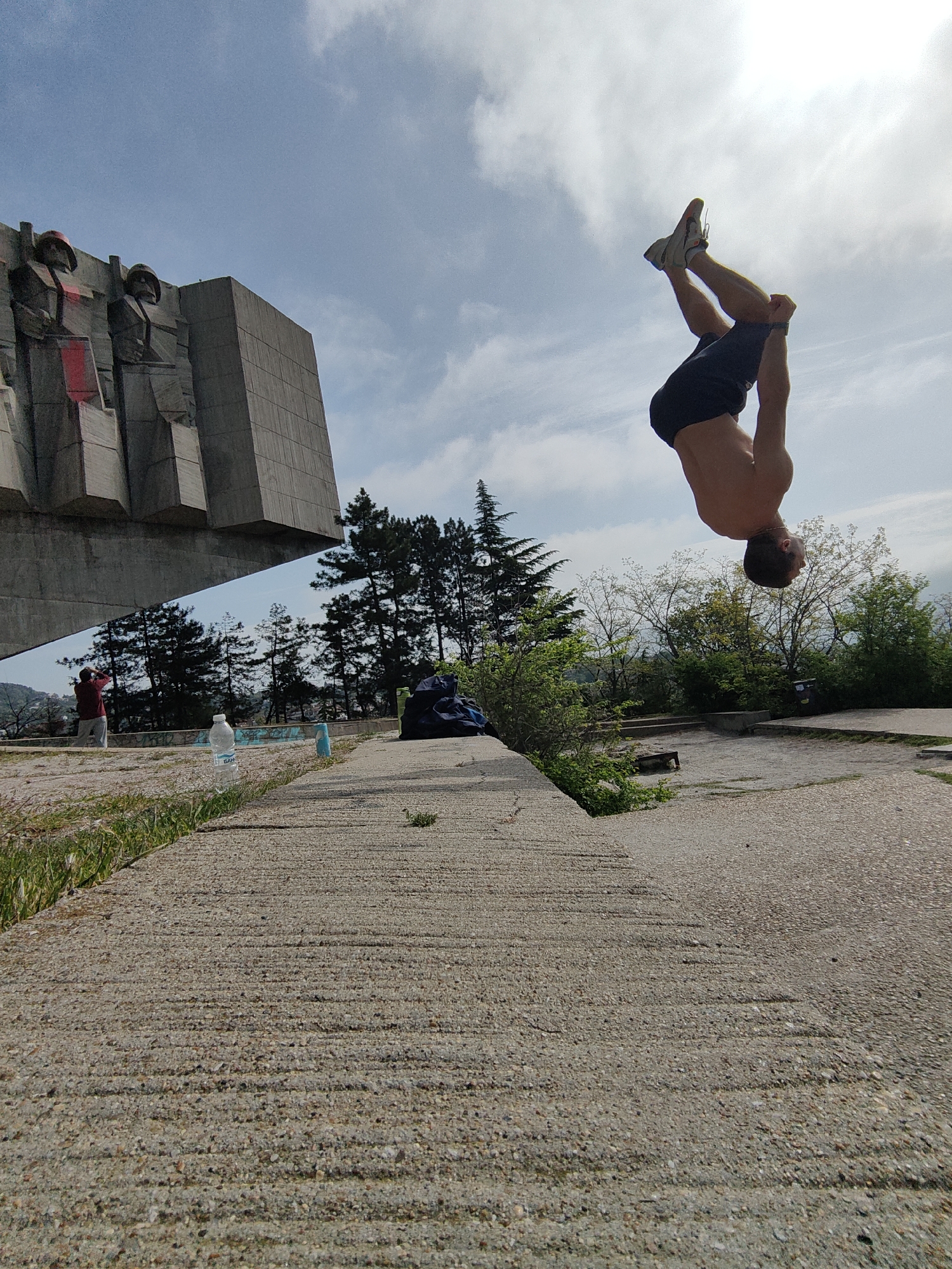 The image captures a man performing a backflip near the Monument of the Bulgarian-Soviet Friendship in Varna, Bulgaria. The monument, a large concrete structure with relief figures, is visible to the left. The man is shirtless, wearing dark shorts and sneakers. He is mid-air, upside down, with his body arched. The surrounding environment consists of a concrete walkway, grass, trees, and a blue sky with scattered clouds. In the background, there are items like a water bottle and a bag, along with a person standing in the distance, taking pictures.