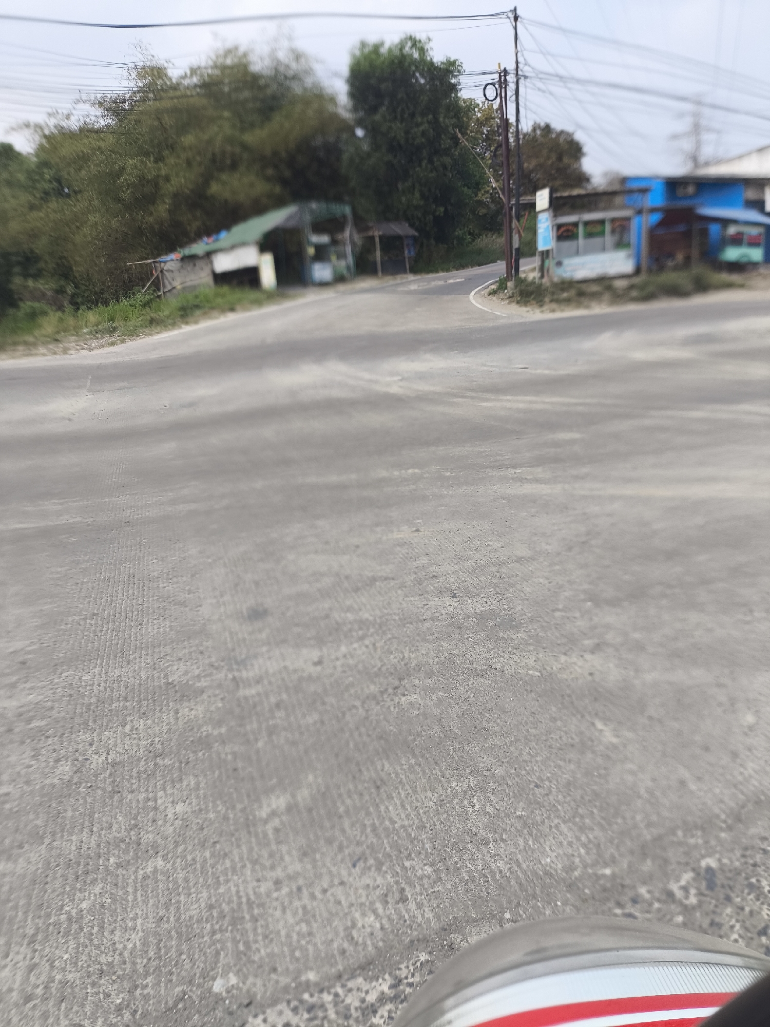 The image shows a road intersection with small roadside businesses. The road is paved with asphalt and appears to be in a rural or semi-urban area. There are several small structures along the side of the road, some of which appear to be shops or stalls. One of the buildings is painted blue and has windows. There are trees and vegetation in the background. Power lines are visible overhead. The scene appears to be in Cilegon, Indonesia, as indicated by the overall environment and typical roadside structures found in that region. The perspective suggests the photo was taken from a low angle, possibly from a vehicle. There are no people visible in the image.