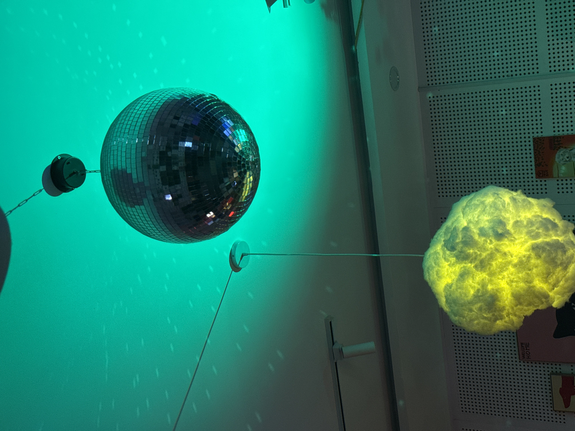 The image shows a disco ball hanging from the ceiling with a light reflecting off of it. Next to the disco ball is a fluffy, glowing object that looks like a cloud. The background is a green wall. The location is not specified, so I do not know the location in the city of Đà Nẵng, Vietnam.