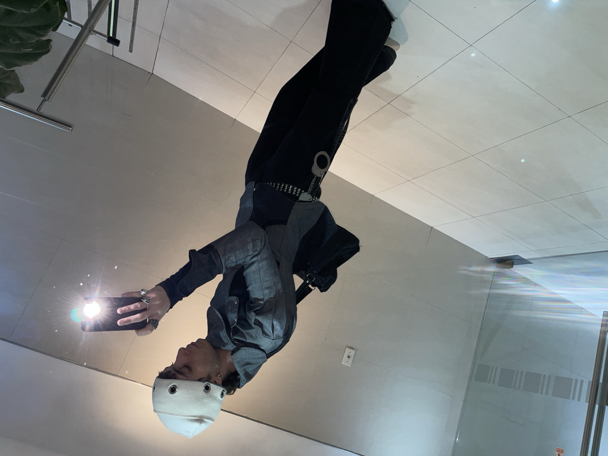 A person wearing a white cap with circular metal eyelets, a gray collared shirt, a black shirt, black pants with a chain and a silver pair of handcuffs attached to the chain, and black shoes is taking a selfie in a location with a light tiled floor. The location seems to be a hallway with a white wall and an electrical outlet on the right side of the person, and what appears to be a glass wall is on the left of the person. The location cannot be determined to be in the city of Buenos Aires, Argentina.