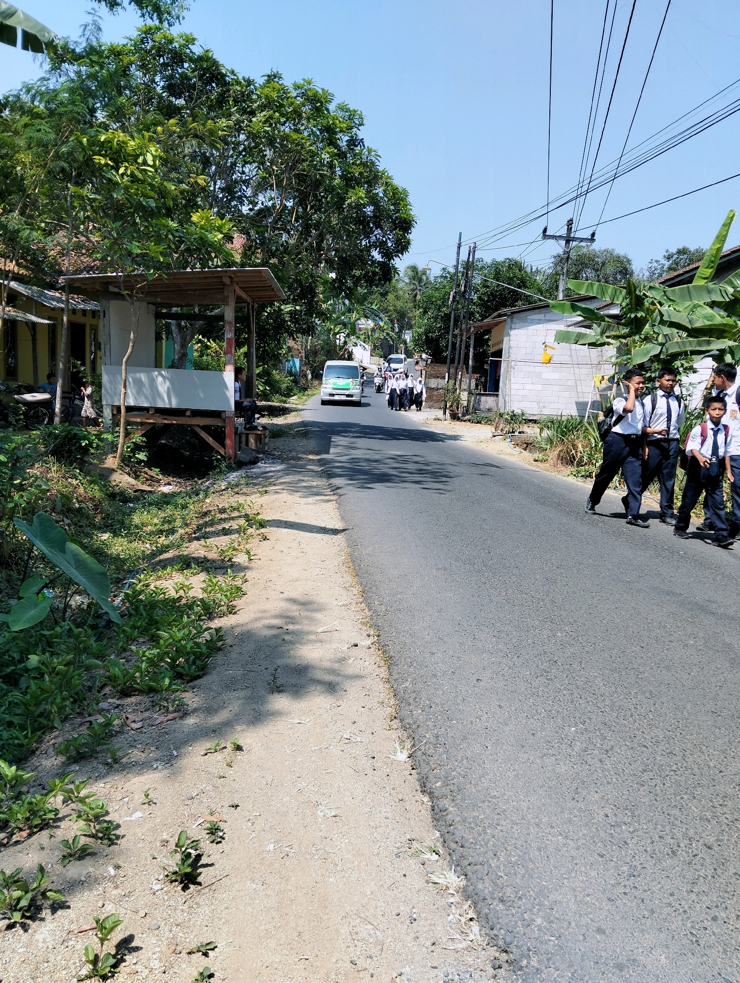 A sunny, clear day in Sidamulih, Indonesia, features a group of Indonesian students in school uniforms walking down a paved road. The students, consisting of boys and girls, are dressed in white shirts and dark pants or skirts, some carrying backpacks. They are walking towards the viewer on the right side of the road. On the left side of the road, a dirt path is lined with green vegetation and a small, open-air shelter with a wooden frame and a metal roof. Further down the road, a white van with a green stripe is visible, along with more students walking. Trees line both sides of the road, and power lines are strung overhead. The scene appears to be a rural or semi-rural area with modest buildings visible in the background.