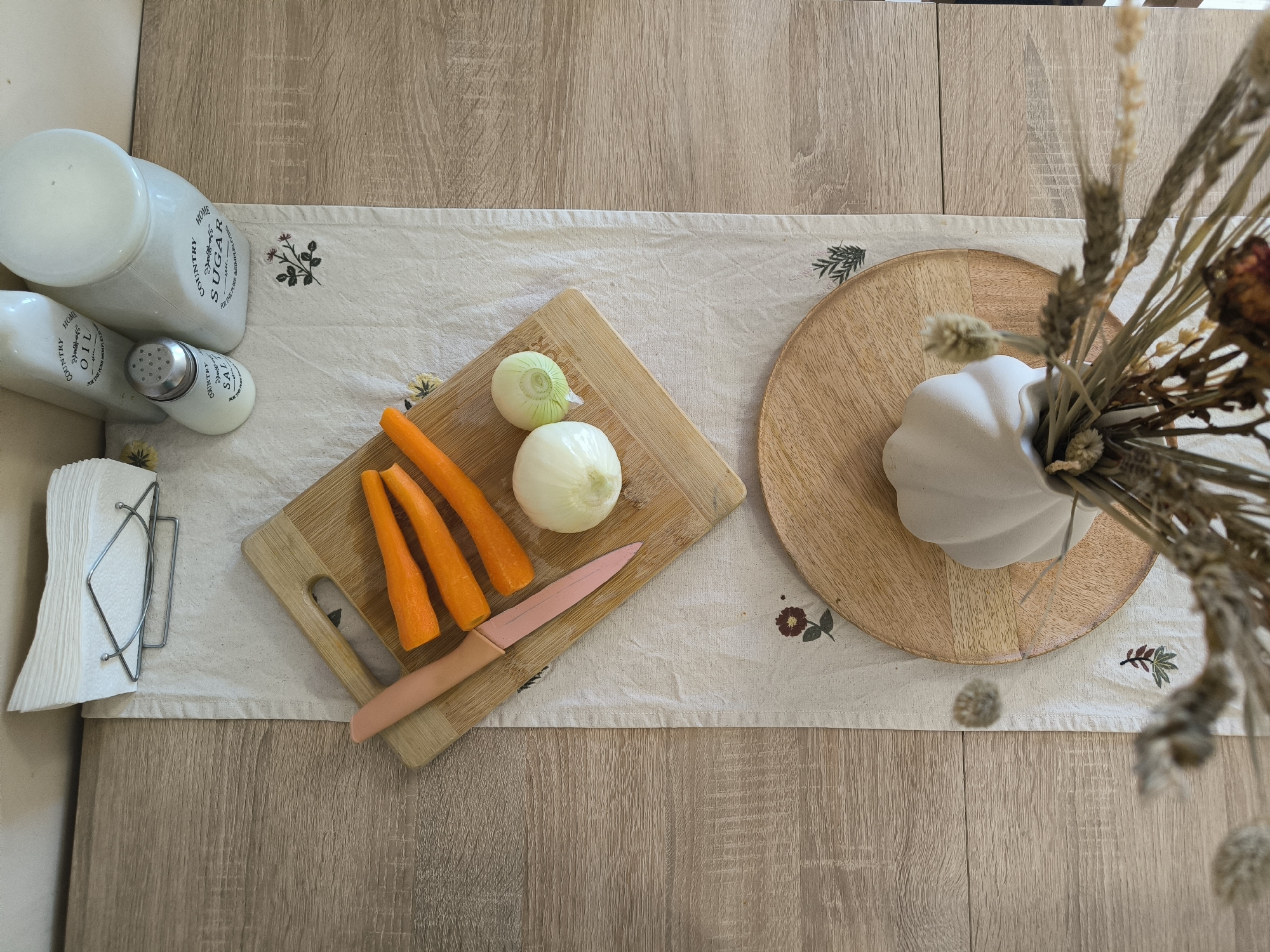 An overhead shot captures a domestic scene on a light wood table, likely in a kitchen or dining area. A cream-colored table runner, embroidered with small floral and leaf motifs, stretches across the table.
In the center, a light wood cutting board holds three peeled orange carrots, a large white onion, and a smaller, light green onion. A pink-handled knife rests on the cutting board beside the vegetables, suggesting food preparation is underway.
To the left, a set of white ceramic or glass containers stands on the table runner. These include a tall cylindrical jar labeled "SUGAR" and "COUNTRY HOME", with additional small text "FOR THE TRUE HOME LOVER" and "GLUTEN FREE". Next to it is an "OIL" bottle and a smaller "SALT" shaker, both also marked with "COUNTRY HOME" and the smaller text. A metal napkin holder filled with white paper napkins is placed to the far left.
To the right, a round wooden placemat or tray holds a white, pumpkin or gourd-shaped vase. This vase contains a rustic arrangement of dried plants, including prominent wheat stalks and other dried flowers or grasses, contributing to a natural or autumnal aesthetic.
The lighting is bright and even, indicating daytime. No individuals are present in the image. The scene presents a functional and decorative arrangement, highlighting kitchen essentials and natural accents.