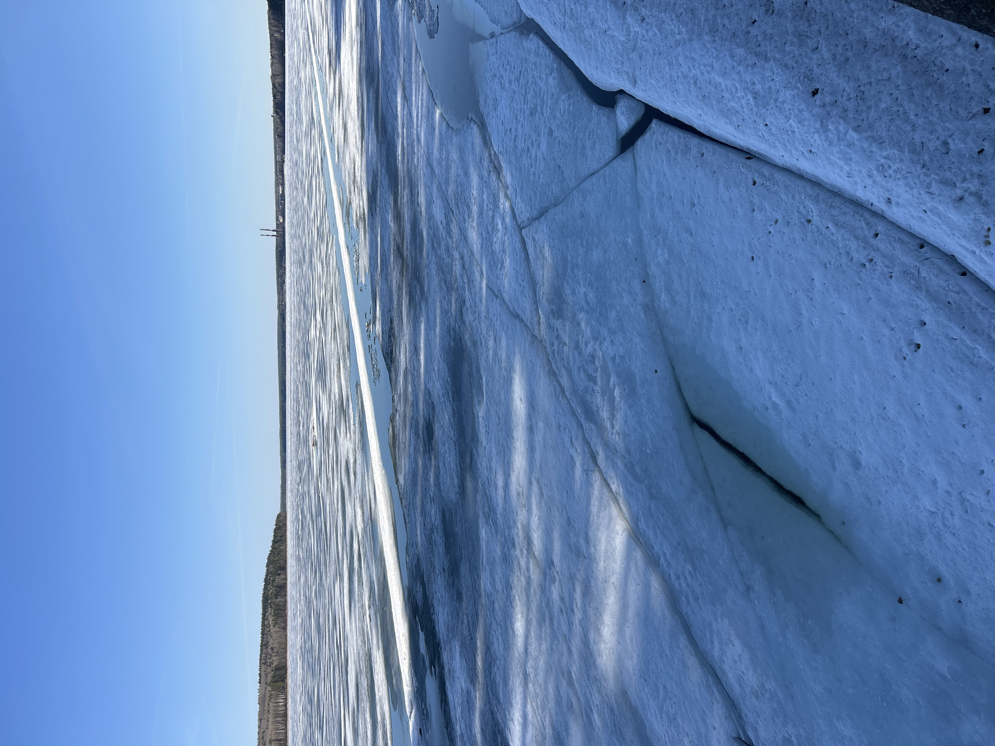 This media file captures a vast expanse of ice-covered water, likely a large lake or river, under a clear blue sky, suggesting a bright sunny day. The scene depicts the process of ice breaking up, characteristic of a spring thaw.
In the foreground and midground, large, irregular sheets of blue-white ice dominate the frame. These ice sheets are fractured with prominent dark cracks, and in some areas, open water is visible between the broken chunks. The surface of the ice appears textured and slightly granular, indicating melting. Darker specks, possibly small debris or dirt, are scattered on the ice. Long, narrow shadows are cast across the ice, suggesting sunlight from a particular angle, likely mid-morning or afternoon.
The horizon features a distant, dark green treeline stretching across the image, marking the opposite shore. Above the treeline, on the left side, two tall, slender industrial structures, possibly chimneys, are discernible against the sky. The sky above is a uniform, clear blue, without clouds.
No people or explicit activities are visible, emphasizing the natural event of the ice breaking up. There is no visible text in the image. The setting aligns with the provided context of Chaikovsky, Russia, where such an environment during seasonal changes is common.