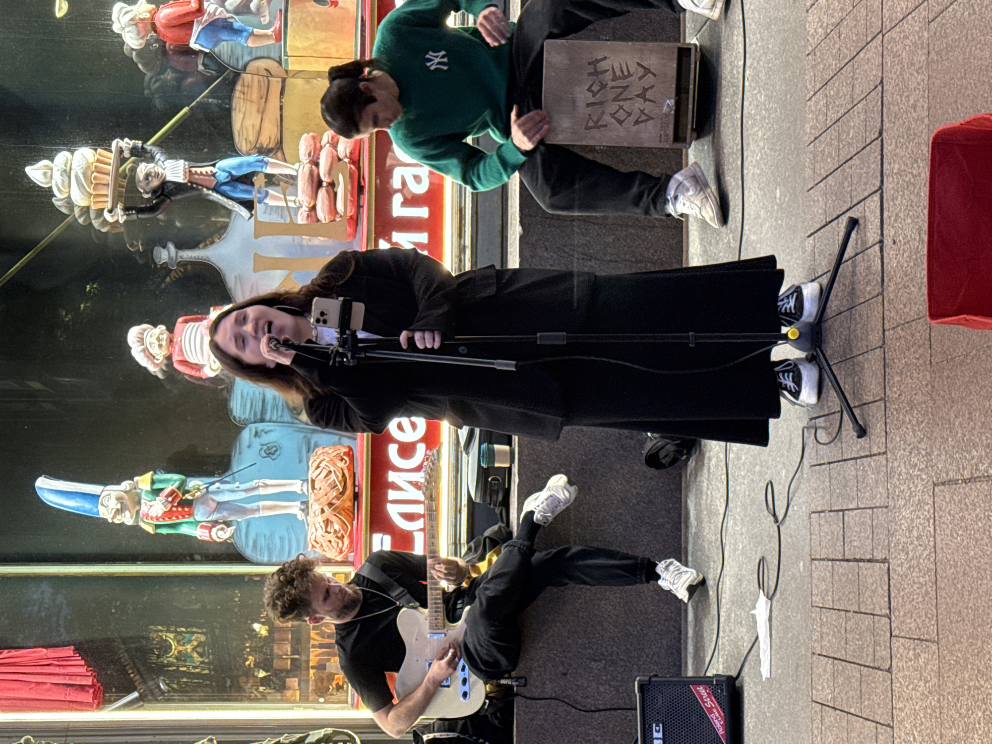 The image depicts a street performance in front of a storefront window. A woman is singing into a microphone, holding a smartphone on a stand. To her left, a man is playing an electric guitar while sitting on a ledge. To her right, another man is sitting on a wooden box, playing it as a percussion instrument. The storefront window displays several animated figures, including a nutcracker-like soldier and a figure holding a large cupcake. Text in Russian is visible on the storefront window. The performers are on a paved sidewalk. The overall scene suggests a lively urban environment.