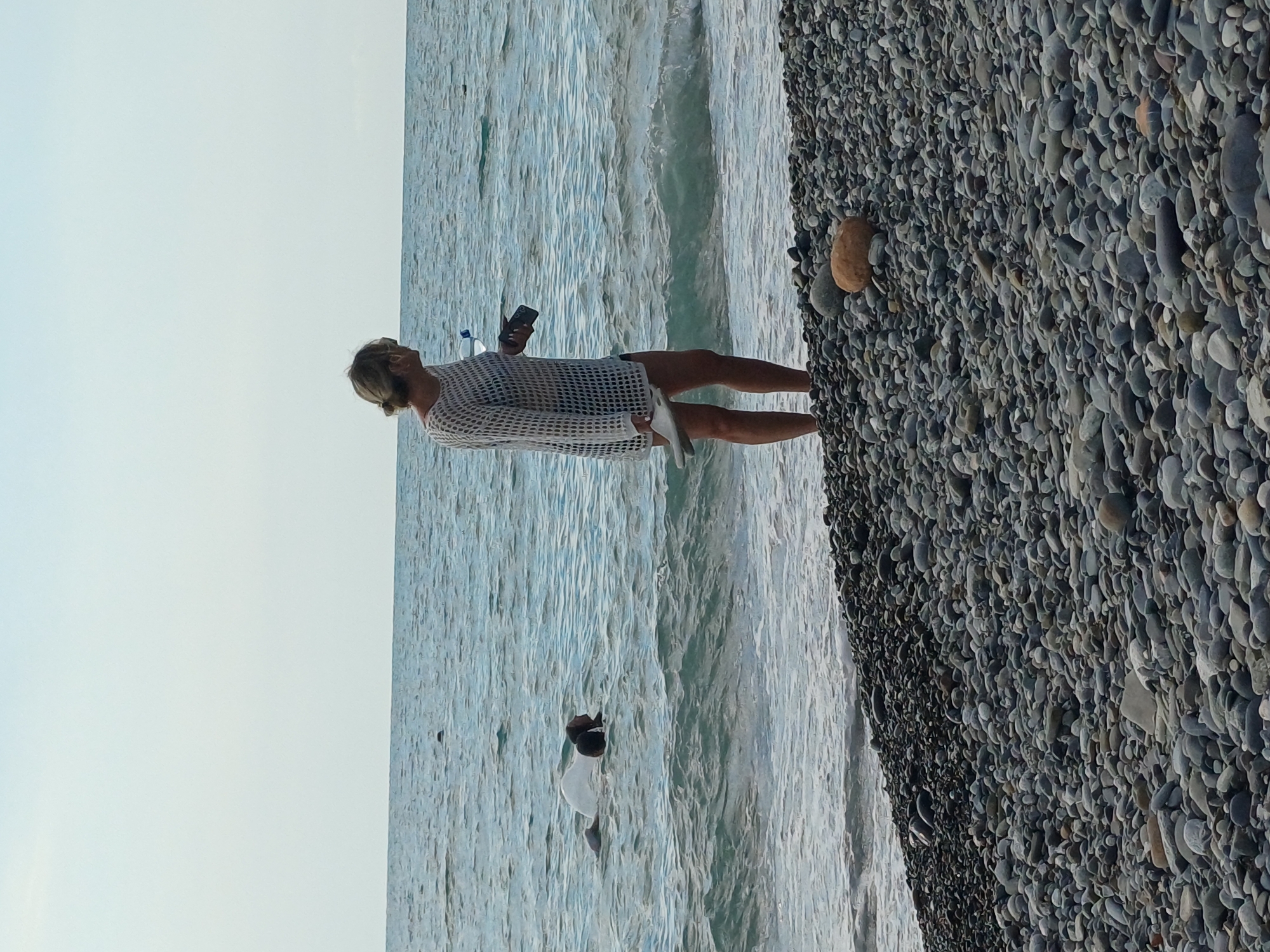 A woman stands on a pebble beach facing the ocean. She is wearing a white mesh cover-up over black shorts, and holding a smartphone and a clear plastic water bottle. In the water, a person is partially submerged, with a white object, possibly a bag, floating nearby. The sea is calm with small waves. The sky is overcast. The image appears to have been taken in Sochi, Russia.