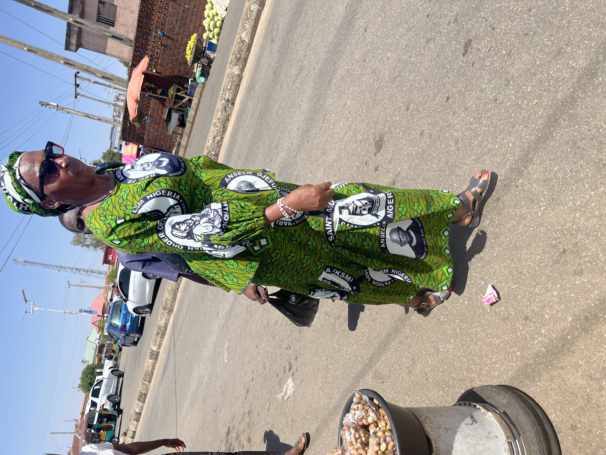 A street scene is dominated by a woman wearing a green and white patterned dress featuring the name "ANSELM OJEJUFHOOL". The dress also has repeating images of a man's face alongside the words "LSM NIGERIA", "SAINT MU", and "PATRON SAINT". She has a matching head wrap and dark sunglasses. She is standing on a paved road with cars and buildings visible in the background. A container filled with packaged items is in the bottom-right of the frame. Some trash is visible on the ground.