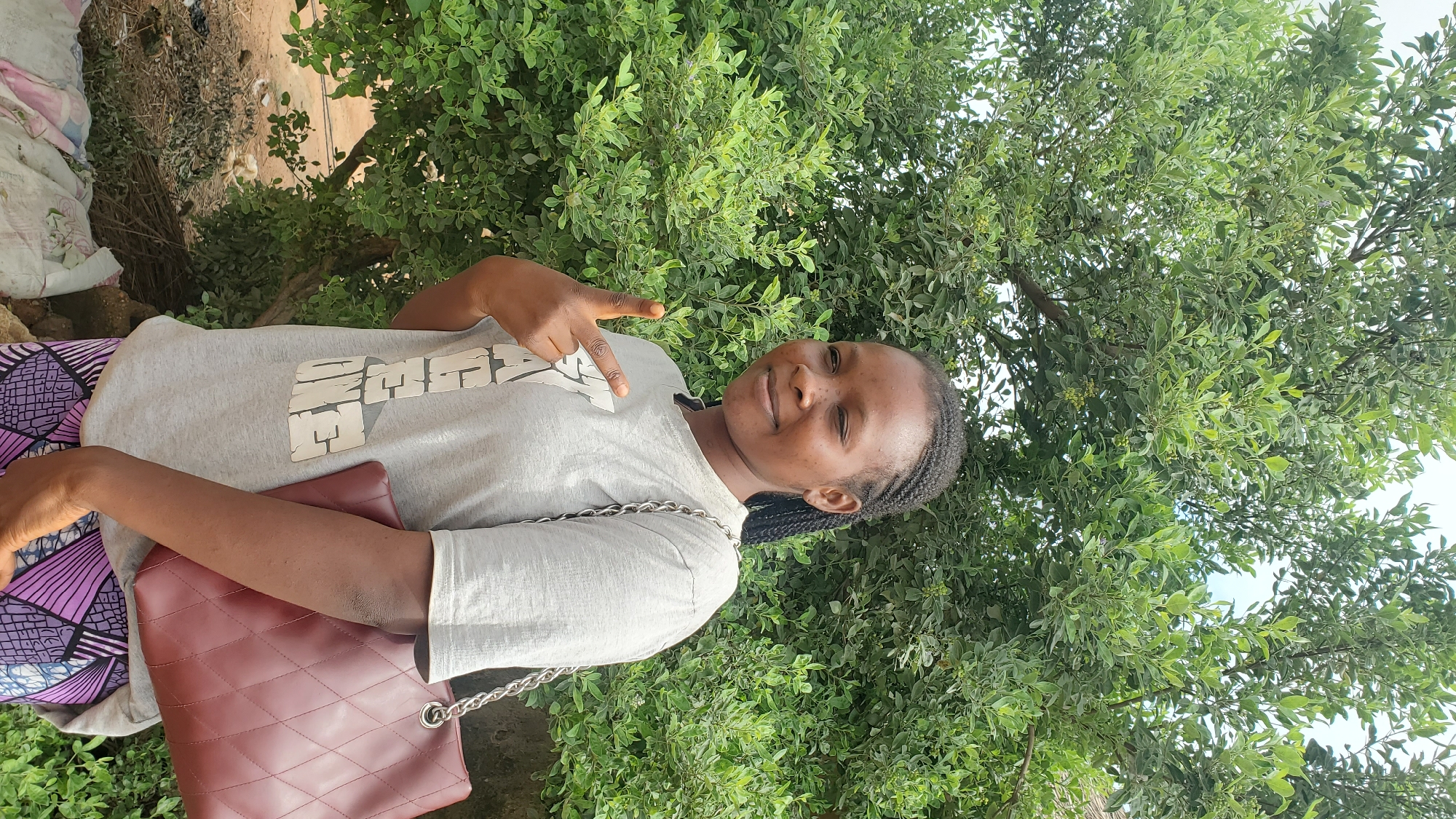 A young Black female with dark braided hair, a light grey t-shirt with a graphic print, and a patterned skirt stands outdoors, smiling and winking with her left eye while making a peace sign with her right hand. She is holding a quilted burgundy purse with a silver chain strap. The background consists of lush green foliage from a large tree. The ground beneath her feet is covered with dirt and scattered debris, including some fabric. The setting appears to be a residential area or a public space with natural vegetation in Jalingo, Nigeria.