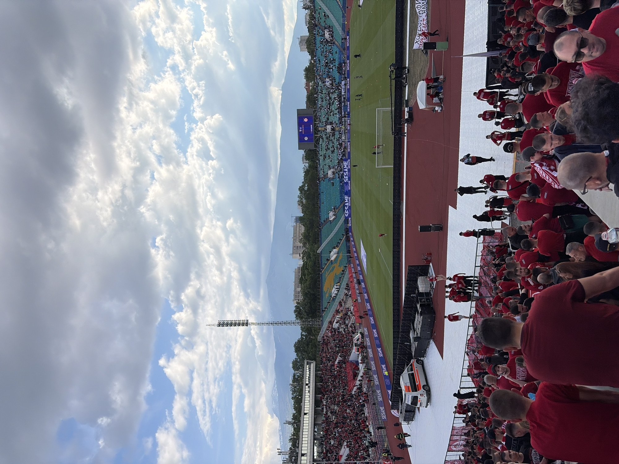Here is a description of the image:
The image shows a soccer stadium filled with fans, likely located in Sofia, Bulgaria, based on the mountain range and building style. The sky above has a mix of clear blue and overcast conditions. The stadium has an active soccer match in progress, and the stands are densely packed with supporters. Most of the fans near the camera are wearing red, suggesting they support one of the teams. There are other fans in the stadium. The stadium perimeter has advertisement banners along the rails. An ambulance is parked near the stands. A high-mast lighting system is also present.