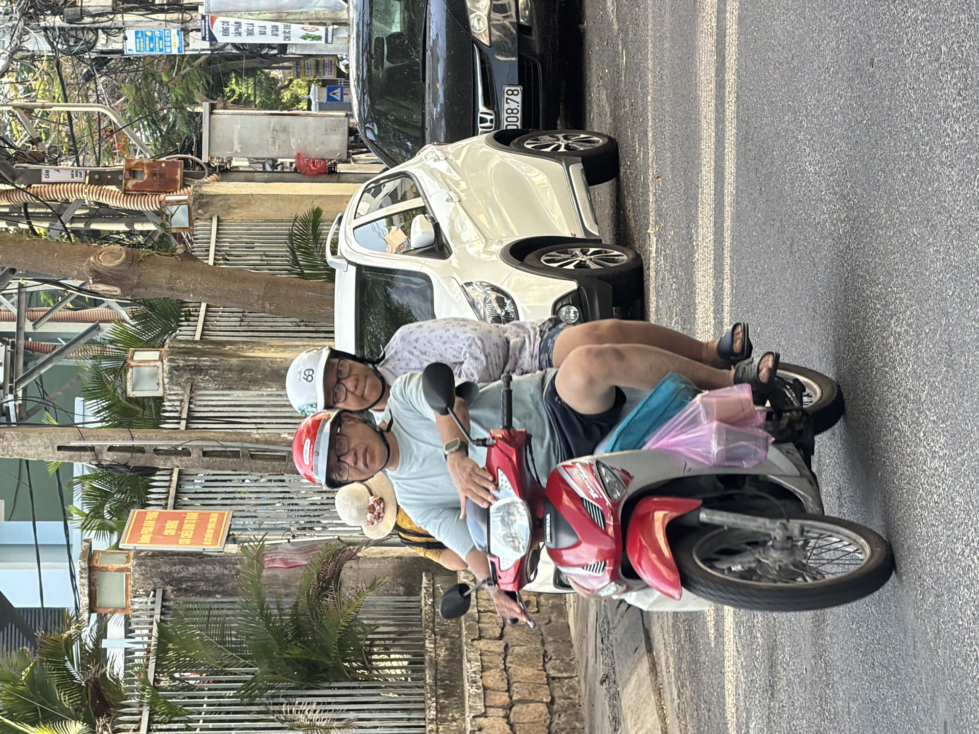A man wearing a red helmet and a light blue t-shirt, along with a woman wearing a white helmet and a patterned shirt, are riding a red and silver motorcycle on a city street in Nha Trang, Vietnam. The man is driving, with his hands on the handlebars. The woman is seated behind him. They are both wearing glasses. A third person, wearing a straw hat, is partially visible on the back of the motorcycle, wearing a yellow top. A pink plastic bag with containers is hanging from the side of the motorcycle. A white car is to the right of the motorcycle, followed by a black car. The background shows a stone wall with a fence, palm trees, and utility poles with numerous electrical wires. A red and yellow sign with text is mounted on the fence. The street surface is asphalt with white lane markings. The scene appears to be during daylight, with sunlight casting shadows.