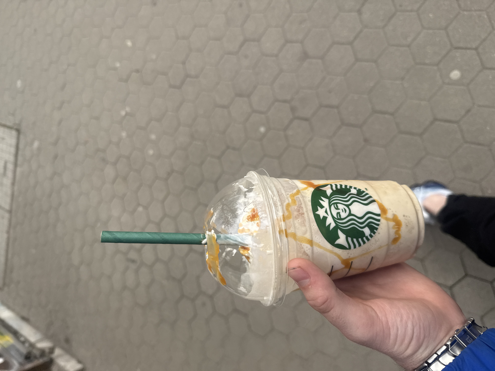 The image captures a person's hand holding a nearly empty Starbucks Frappuccino cup outdoors in Sofia, Bulgaria. The cup is made of clear plastic with a domed lid, showcasing the remaining light-colored beverage and distinct streaks of caramel sauce both inside and on the exterior of the lid. A green paper straw is inserted into the lid, slightly bent where it enters. The prominent green and white Starbucks siren logo is visible on the cup's side.
The hand, with visible dirt or stain under the fingernails, belongs to a person wearing a metal-banded watch on their wrist and a blue sleeve. In the background, a gray paved surface with a distinctive hexagonal tile pattern extends, suggesting an urban outdoor environment like a sidewalk or plaza. A glimpse of another individual's foot, clad in a white and blue athletic shoe and black trousers, is seen in the upper right. The scene is brightly lit, indicating daytime. Small, faint text, possibly "RECYCLE ME," can be discerned on the clear plastic lid.