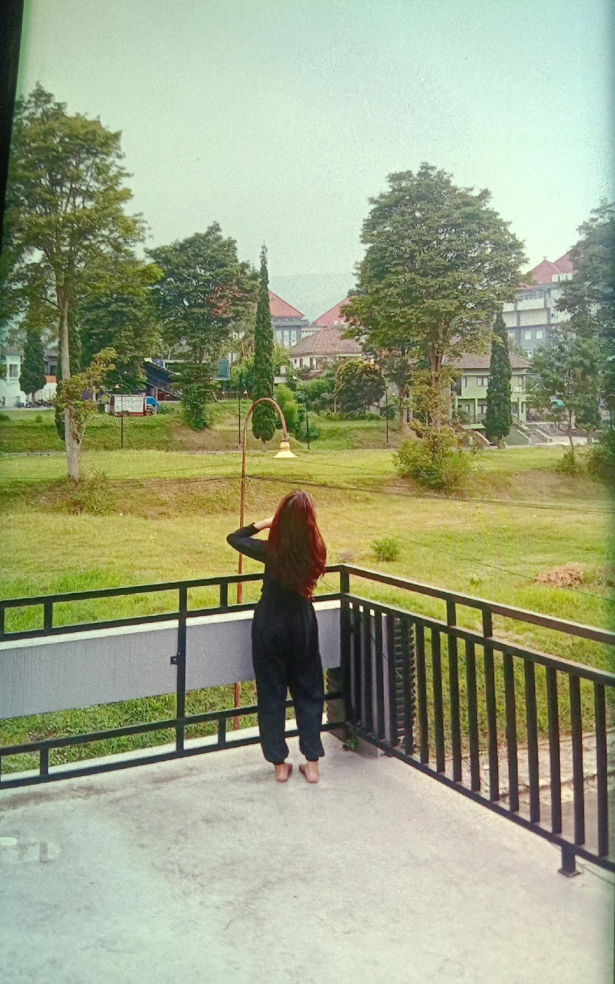 A young woman stands with her back to the camera on a concrete balcony, looking out over a grassy area with trees and buildings in the background. She has long, reddish-brown hair and is wearing black, loose-fitting pants and no shoes. Her right arm is bent at the elbow, with her hand near the back of her head, and her left arm is down by her side. The balcony has a grey railing with black metal bars. Beyond the balcony, a large expanse of green grass is visible, dotted with trees of various sizes. In the middle ground, there is a decorative lamppost with a yellow lamp. Further in the background, several buildings with red-tiled roofs and white facades are nestled amongst more trees. The sky is a pale, hazy blue, suggesting an overcast day. The overall setting appears to be a university campus or a similar institutional environment, likely in Gampengrejo, Indonesia, given the lush greenery and architectural style.