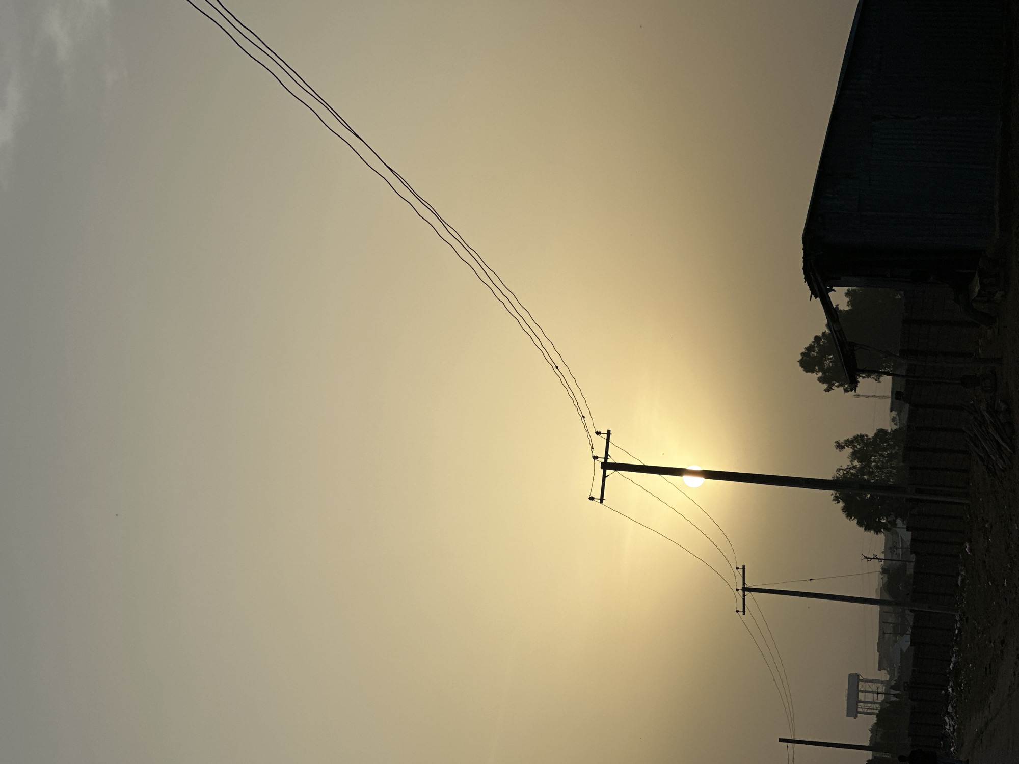 This vertically oriented image captures a hazy outdoor scene during either sunrise or sunset, characterized by soft, diffused lighting. A bright, circular sun, low in the sky, is the primary light source, partially obscured by utility poles and power lines on the upper right side of the frame. The sky exhibits a warm yellow-orange glow around the sun, gradually fading into a dull grey-yellow and light grey further away, with faint clouds visible in the upper left.
The foreground and mid-ground are dominated by the dark silhouettes of multiple utility poles with cross arms and numerous power lines stretching diagonally across the sky from the lower left to the upper right. On the far right side of the image, a large, dark, industrial-looking building with a sloped corrugated metal roof is visible, along with silhouetted trees and dense vegetation. In the hazy distance on the right, the outline of a water tower and other faint structures can be discerned. The immediate foreground on the right appears to be an unpaved dirt road. The overall impression is one of a quiet, twilight landscape, possibly experiencing dusty or hazy weather conditions. No people or visible text are present. The scene is consistent with settings found in Bukuru, Nigeria.