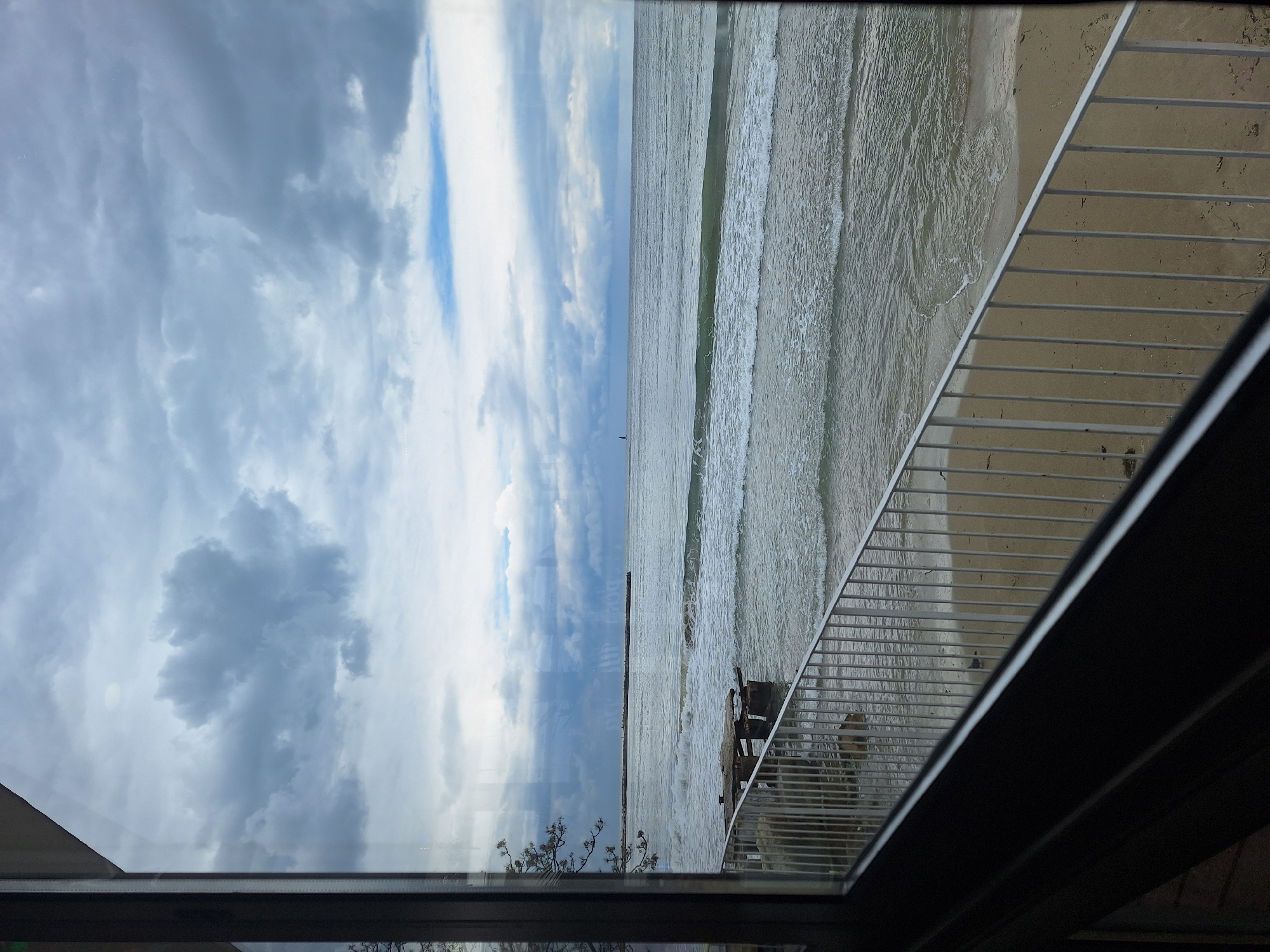 The image shows a view of the Black Sea coastline in Varna, Bulgaria, seen through a window. The sky is mostly overcast with patches of blue. The sea has waves breaking on the sandy beach. A partially submerged pier or breakwater extends into the sea. A metal railing, possibly belonging to a balcony or a staircase, is visible on the right side of the frame. Inside the window, there is a reflection of a tree branch.