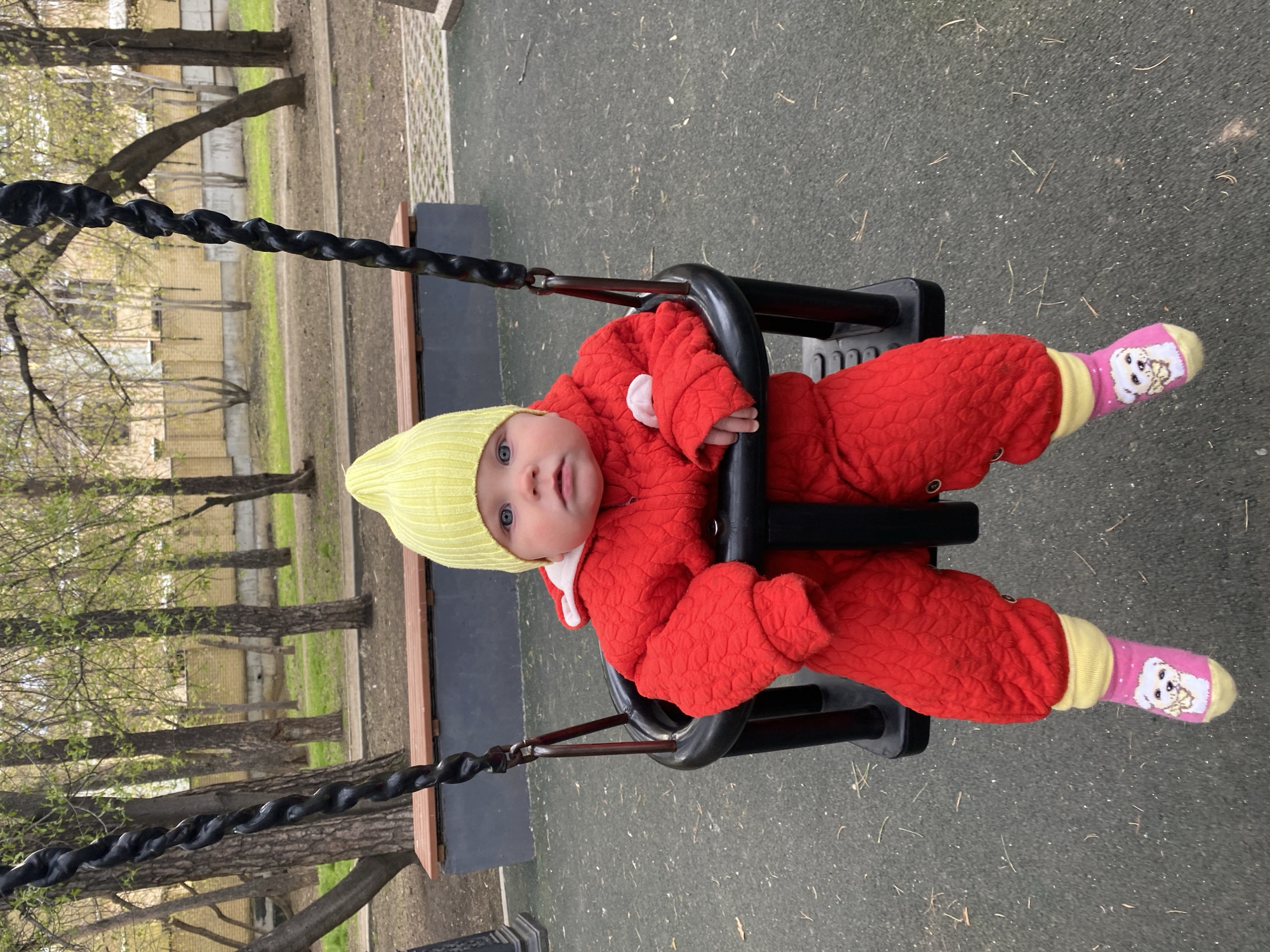 An infant is seen in a close-up, seated in a black plastic bucket swing at a playground in Yekaterinburg, Russia. The baby, with striking light blue eyes, looks directly at the viewer with a calm and curious expression. They are dressed for cool weather in a vibrant red quilted full-body suit with a white collar, and a light yellow knitted beanie. Their feet are adorned with playful pink socks featuring a cartoon white dog wearing glasses and a bow, with yellow cuffs.
The swing is suspended by thick, dark, twisted chains. The ground beneath is a dark grey, speckled rubber safety surface, typical of playgrounds. In the background, bare trees show the initial signs of spring with nascent green leaves, suggesting the season is early spring. A light-colored brick building or fence is visible through the trees in the distance. A wooden bench or raised border is partially seen to the left of the swing. The lighting is natural and soft, indicating daytime, possibly under an overcast sky. There is no visible text in the image.