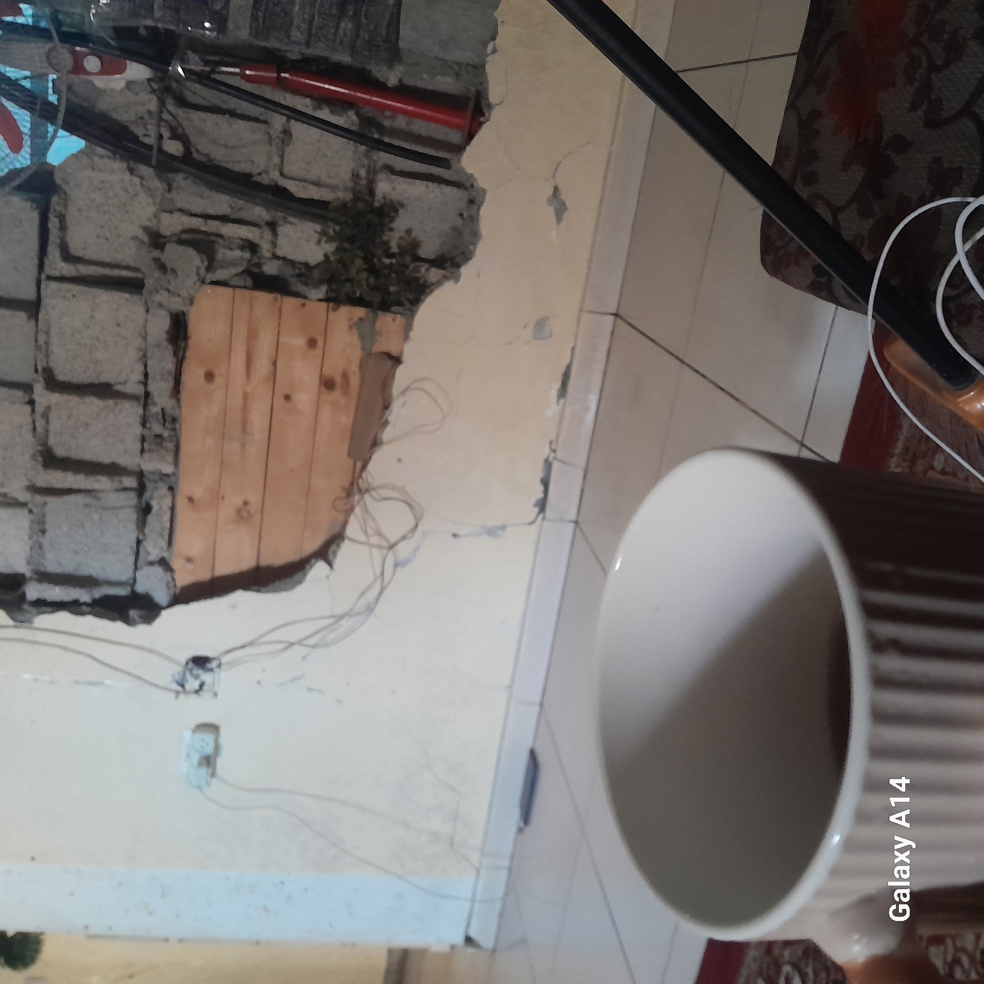 An interior scene in Khan Yunis, Palestinian Territory, depicts a severely damaged room. In the foreground, a light-colored, vertically ribbed mug containing a dark liquid is held by a partially visible hand, positioned over a reddish-brown patterned rug. To the right, a black rod extends diagonally across a patterned cushion, where white electrical cables are loosely coiled. The floor consists of light-colored square tiles with dark grout lines; a blue rectangular object rests on it near the base of the wall.
The wall shows extensive damage: its light-colored plaster is cracked, peeling, and missing in several areas, revealing the underlying structure. A large, irregular breach exposes courses of gray cinder blocks in the upper section of the opening. Below these blocks, a segment of the opening is covered by horizontally arranged wooden planks, suggesting a temporary repair or seal. Dark green vegetation grows directly from the exposed wall cavity to the right of the planks. Loose electrical wires are draped across the damaged plaster, and two electrical outlets are visible, one partially intact, the other damaged with exposed wiring. To the far right, a red cylindrical object and several dark, slender rods are leaning against the compromised wall, while a section of light blue tarp or fabric is partially visible through an upper opening in the structure. The image was captured by a device identified as "Galaxy A14".