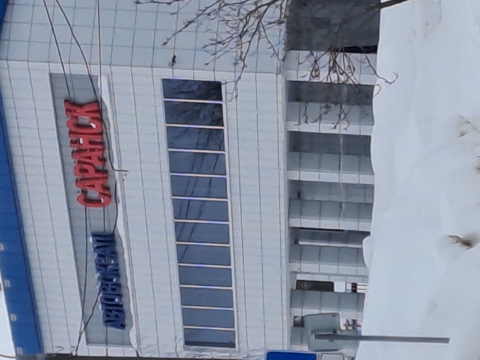 A tilted eye-level shot captures a multi-story building with "CAPAHCK" vertically displayed in red lettering. Beneath it, "АВТОКРАЙ" appears in blue. The building features a line of narrow, tall windows. Adjacent to the building, a gray structure of horizontal levels is visible, partially obscured by a large snowdrift. Bare tree branches are seen above the building.