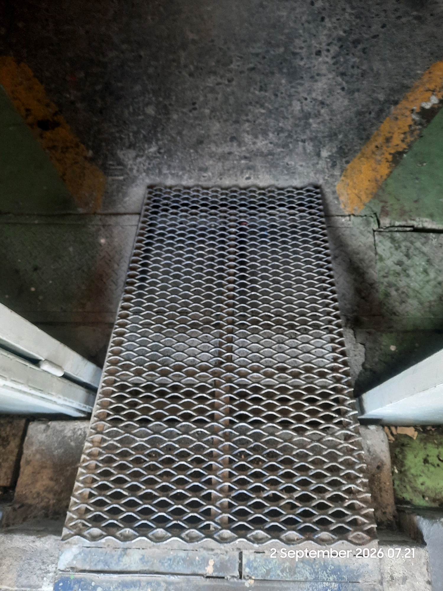 The image shows a metal grate covering a drain, likely in an industrial or utility area. The grate is made of diamond-shaped expanded metal and is positioned horizontally, taking up the majority of the frame. The surrounding environment appears to be concrete and asphalt. There are yellow painted lines on the concrete, indicating some form of demarcation or safety marking. The scene is viewed from above, looking down. The overall impression is one of an outdoor, functional space. The timestamp in the bottom right corner indicates the image was taken on September 2, 2026, at 07:21.