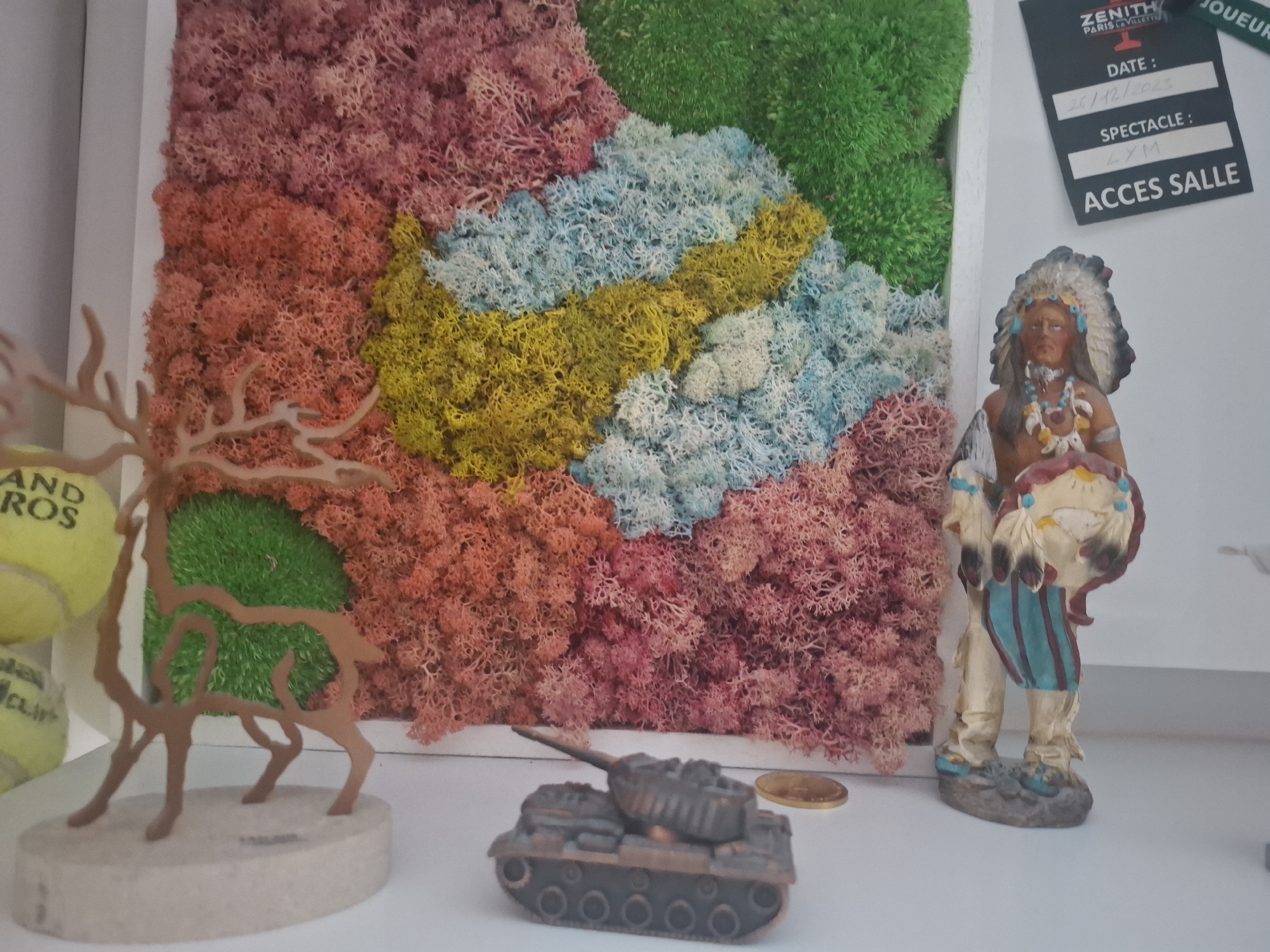 The image depicts a shelf in a room, likely in Montreuil, France, displaying various decorative items. A central feature is a framed artwork composed of colorful dried moss in hues of pink, coral, yellow, light blue, and green, arranged in a textured, abstract pattern. To the left of the moss art is a brown, silhouetted sculpture of a deer. Partially visible to the far left are two yellow tennis balls, one with partial text "AND ROS". In the foreground, a miniature dark grey tank is positioned on the shelf. To the right of the tank is a small gold-colored coin. Further to the right stands a figurine of a Native American man adorned with a feathered headdress, beads, and traditional clothing, holding a shield. Attached to the wall behind the figurine is a dark rectangular card with white text, which appears to be a ticket or pass. The text includes "ZENITH PARIS LA VILLETTE", "DATE : 26/12/2023", "SPECTACLE : LXM", and "ACCES SALLE". The background is a plain white wall.