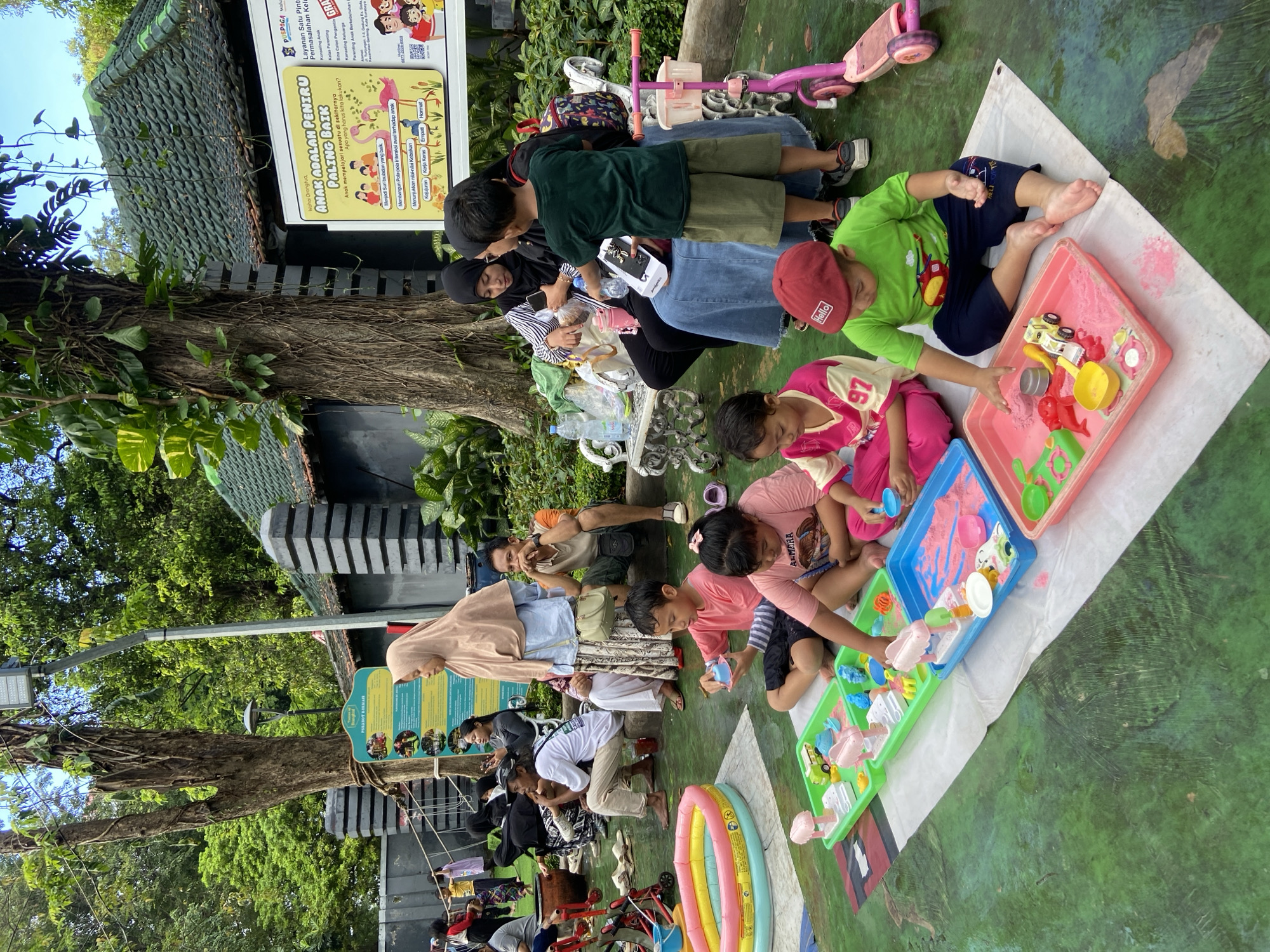 The image captures a scene in what appears to be a public park or recreational area in Surabaya, Indonesia. A group of children are engaged in play, interacting with colorful plastic toys in trays filled with what looks like sand or a similar play material. They are seated on a white cloth spread on the ground, which is marked with a green surface pattern.
Adults, presumably caregivers, are seated or lying nearby, observing and interacting with the children. Some are seated on benches, while others are closer to the children on the ground. There is a variety of personal belongings scattered around, including water bottles, bags, and a small scooter.
In the background, there is a signpost with information written in Indonesian. Trees and other vegetation surround the area, suggesting an outdoor setting. The building structure has a dark-colored exterior with a corrugated roof. The overall atmosphere suggests a family-friendly environment where children are engaged in play under the supervision of adults.