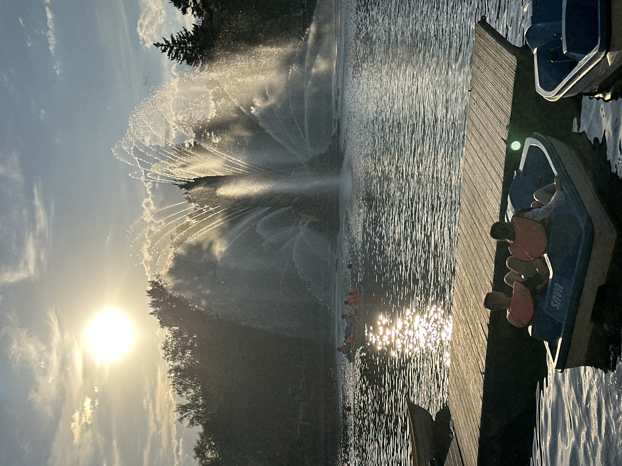 The image displays a picturesque golden hour scene at a lake or large pond in Moscow, Russia, likely within a park. A prominent, multi-tiered fountain dominates the background, spraying numerous jets of water high into the air, which glisten brightly as the low sun illuminates them.
The sun, a brilliant orb, is positioned low in the sky on the left side of the frame, casting strong, sparkling reflections across the rippling surface of the water, which appears dark but reflects bright patches of sunlight. Dark silhouettes of trees line the far bank of the water body, providing a natural backdrop to the fountain.
In the foreground on the right, a wooden dock extends into the water, where several pedal boats are moored. Two individuals, one an adult and one a child, are visible near a blue pedal boat, both wearing orange life vests, suggesting they are either preparing to embark or have just returned from a ride. The word "SAVA" is visible on the side of this boat. Further out on the water, another boat, also carrying several people, can be seen closer to the fountain. The sky is partly cloudy with some wispy formations around the sun. The scene conveys a relaxed, leisure atmosphere during what appears to be a late afternoon or early evening outing.