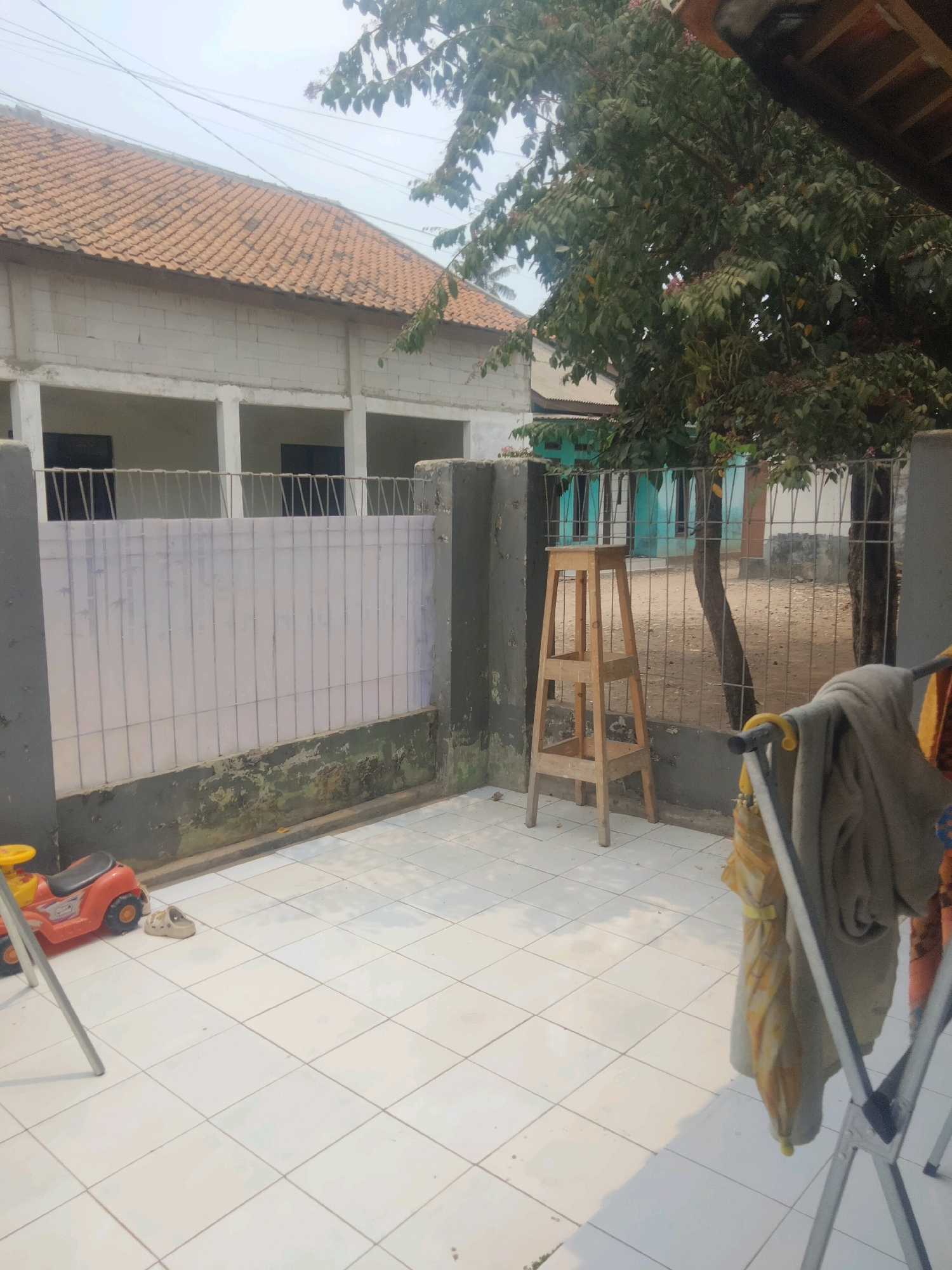 The image captures a residential outdoor area in Suka Sari, Indonesia, on a bright, overcast day. In the foreground, white tiled flooring is visible, reflecting the ambient light. To the left, a small, red and black toy car is positioned, with a pair of light-colored shoes nearby. A metal drying rack with several pieces of clothing hanging on it occupies the right side of the frame, casting a distinct shadow on the tiles. Behind the foreground elements, a gray concrete wall with peeling paint runs horizontally. A metal fence with vertical bars and a white panel is situated in front of the wall, with a portion of the fence showing some discoloration. A wooden step stool with three tiers stands near the fence.
In the mid-ground, behind the fence, is a building constructed with white cinder blocks and topped with reddish-brown roof tiles. This building appears to be a house or a similar structure with open window or doorway spaces. Further in the background, a vibrant turquoise-colored building with windows is visible. A large tree with dense green foliage stands prominently, its branches extending over the scene, partially obscuring a view of the sky. Electrical wires are strung across the sky, suggesting the presence of a power grid. The ground behind the fence appears to be dirt and gravel. The overall scene depicts a somewhat cluttered, yet functional, outdoor space associated with a dwelling in a suburban or semi-urban Indonesian environment.
