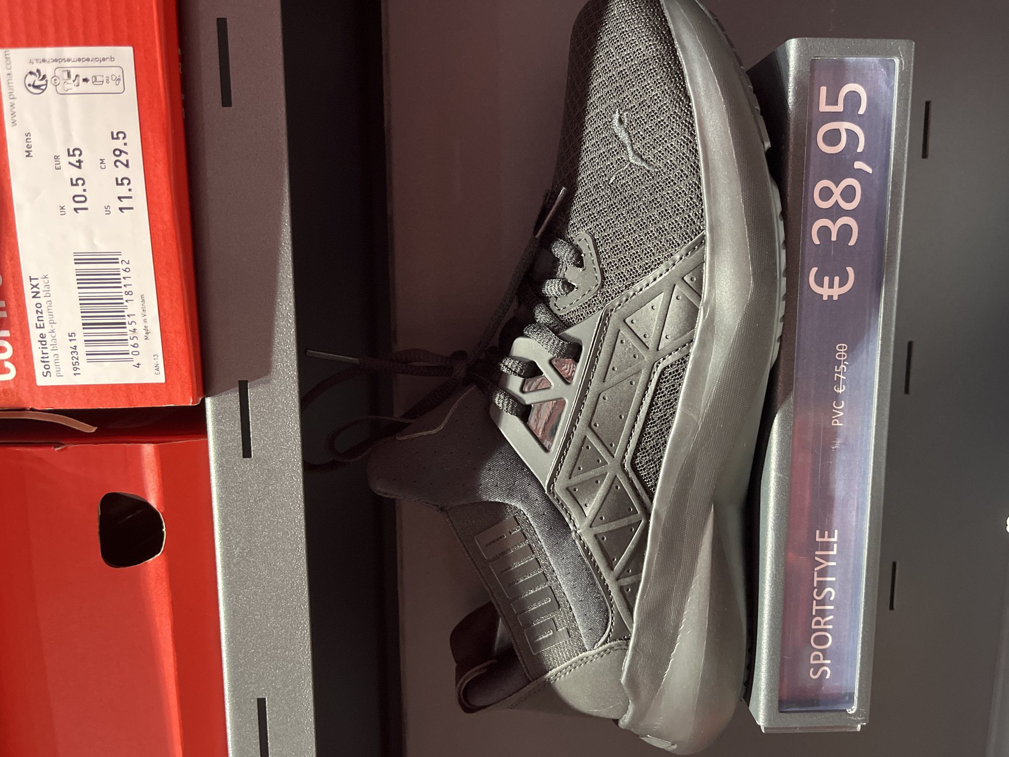 A dark grey Puma Softride Enzo NXT sneaker is displayed on a grey shelf in a retail environment. The sneaker is positioned diagonally, with its toe pointing to the right. A price tag in front of the shoe displays "SPORTSTYLE" and the price "€38,95". Behind the shoe, a red Puma shoebox with a label indicating size information is visible. The label shows "Softride Enzo NXT puma black-pumapuma black" and various size conversions, including UK 10.5, EUR 45, US 11.5, and CM 29.5. The background consists of a dark, blurred surface, likely shelves or walls of a store. The lighting is even, suggesting indoor retail illumination. The scene is static and does not contain any people. This retail display is located in Rodengo, Italy.
