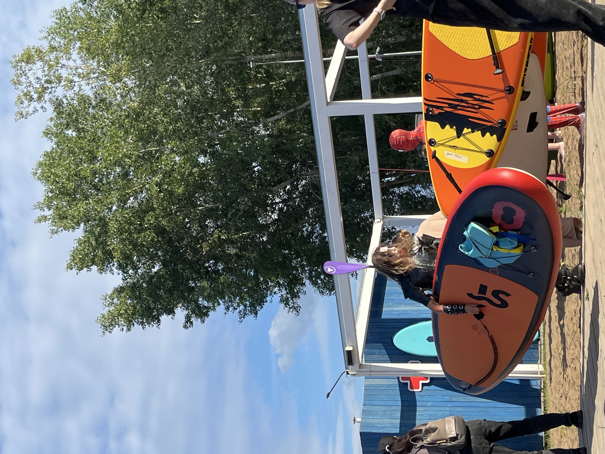 In Chaikovsky, Russia, on a sunny day, several people are gathered on a sandy area near a body of water, possibly a beach or a riverbank. Two individuals are holding stand-up paddleboards. One person, dressed in a black leather-like outfit with dark makeup, is holding an orange and black paddleboard with a yellow pattern. Another person, dressed in a full Spider-Man costume, is also standing near paddleboards. In the background, a blue wooden structure with white framing stands, and lush green trees are visible against a sky with scattered clouds. A person wearing a backpack is standing to the left. The overall scene suggests an outdoor recreational activity.