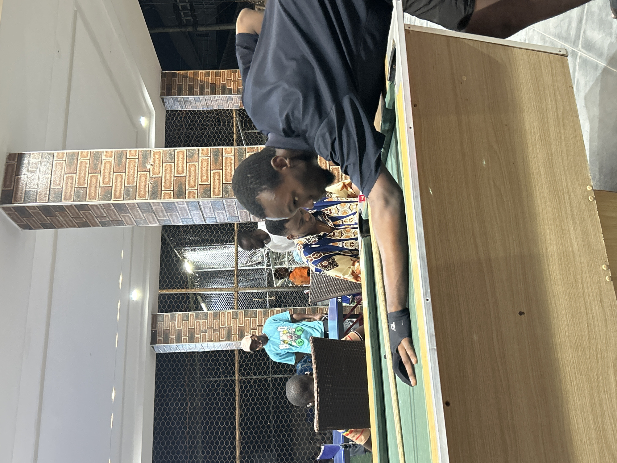 A group of men are gathered around a pool table in an outdoor, covered area, likely in Ihiagwa, Nigeria. The focus is on a man in the foreground, leaning over the pool table, preparing to take a shot. He is wearing a black short-sleeved shirt and a black wristband on his left hand. His right hand is on the pool cue, which rests on the green felt of the table.
In the background, other men are visible. One man in a light blue t-shirt with graphics is standing and watching. Another man in a patterned shirt is seated at the table. Further back, two other individuals are partially obscured. The environment appears to be a recreational space, with a mesh fence in the background and brick-patterned pillars supporting the roof. The lighting suggests it is evening or nighttime. The pool table itself has a wooden body and a metal trim around the playing surface. Other elements visible include chairs and a glimpse of a blue object, possibly another table or chair, to the left. The overall impression is a casual social gathering centered around playing pool.