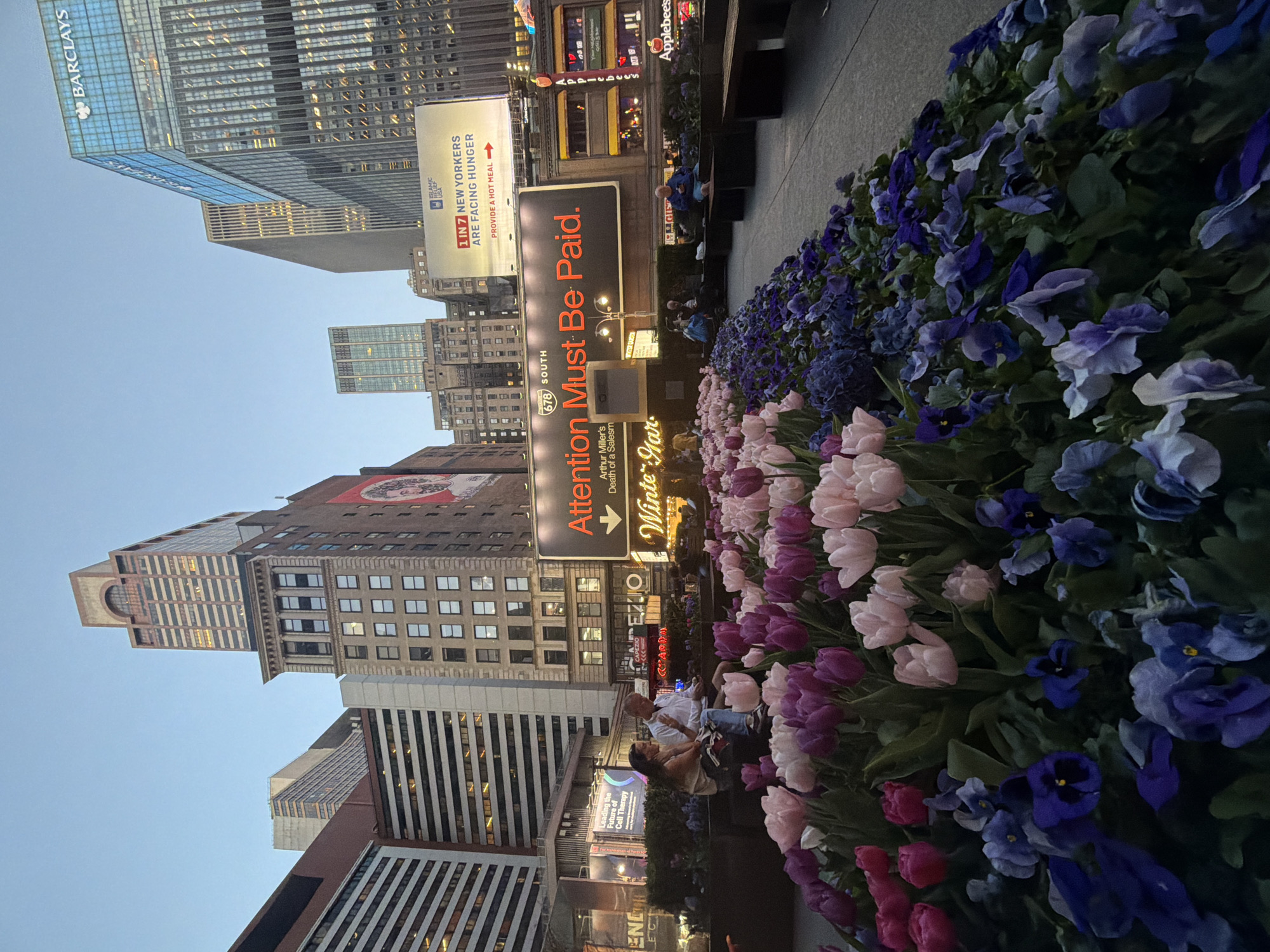 The image depicts a street scene in New York City, with a focus on a flower bed containing a mix of tulips and pansies in shades of pink, purple, and blue. Buildings loom in the background, including the Barclays building, along with billboards advertising cell therapy, Islamic relief, and a play titled "Attention Must Be Paid." A man and a woman are seated on a bench amidst the flowers.