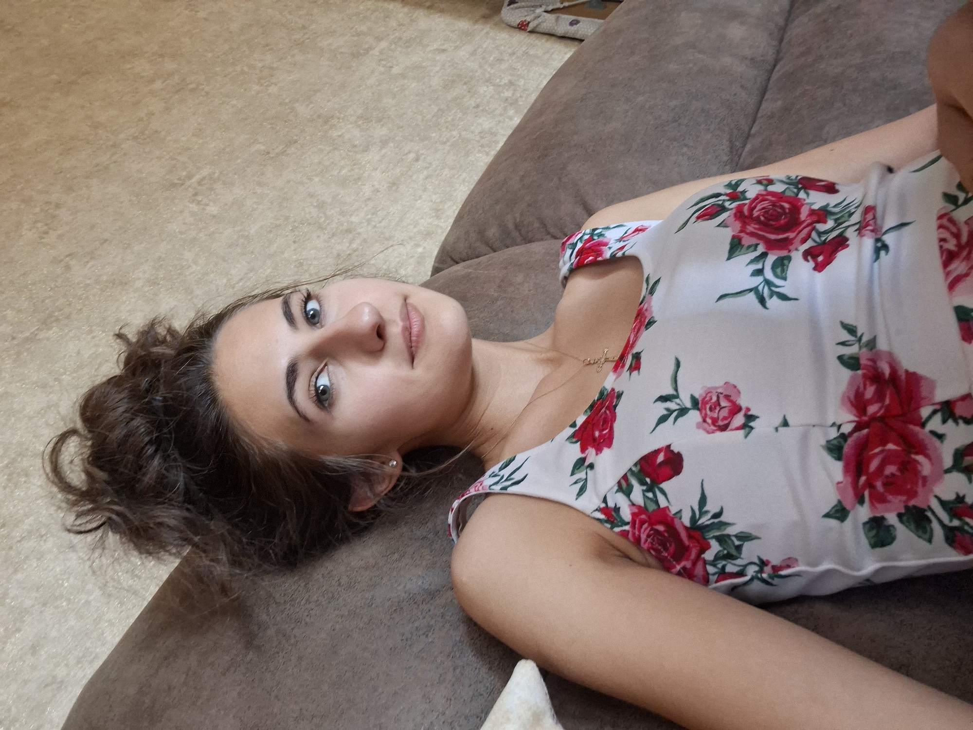 In an indoor setting, a young woman with her hair pulled up in a messy bun reclines on a brown couch. She is wearing a white dress adorned with red roses. Her gaze is directed towards the camera, and she is wearing a delicate gold necklace with a cross pendant. The background wall is textured and beige. The lighting appears to be natural, casting soft shadows.