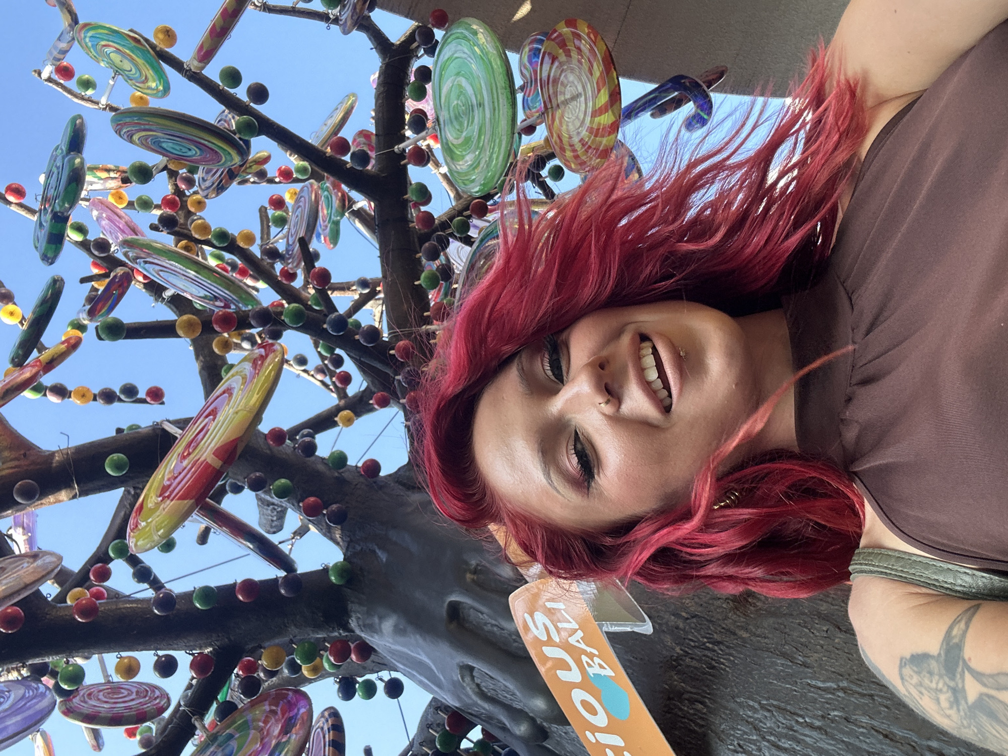 A person with vibrant red hair stands beside a decorative tree with glass lollipops and round balls, likely in Kuta, Indonesia. The individual is positioned close to the camera, smiling, and wearing a brown top. Their left arm, featuring a tattoo, is visible. The foreground reveals a sign, partially visible, that says "licious BALL." The backdrop includes a clear blue sky and segments of the decorative tree laden with colorful glass ornaments.