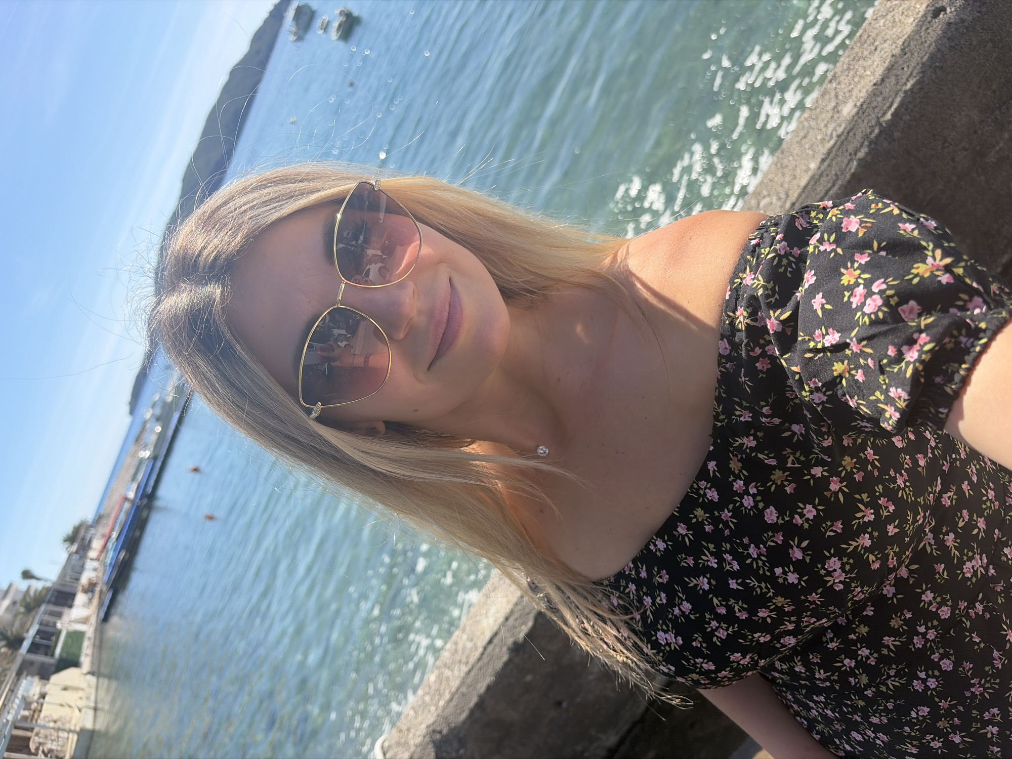 A woman poses in a selfie with a view of the Adriatic Sea in Rafailovići, Montenegro. She is wearing sunglasses and a floral print dress. The sea is visible in the background, with sunlight reflecting off the water. Boats are moored in the distance. A concrete structure is in the foreground.