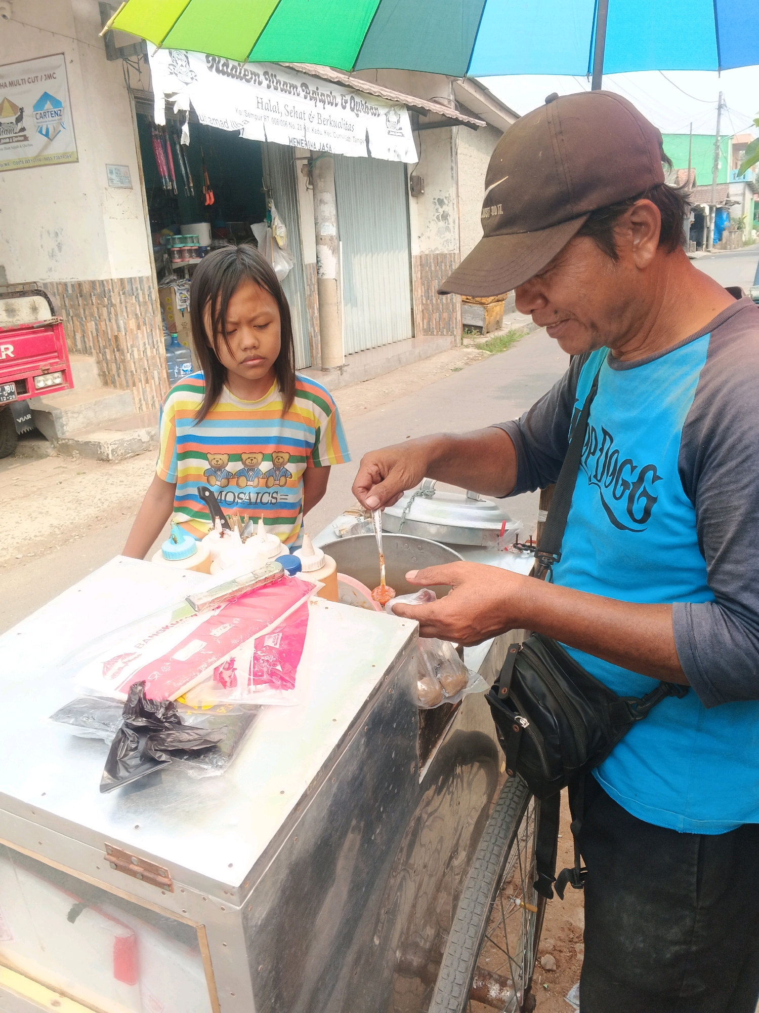 A man in a brown baseball cap and a blue and gray t-shirt stands behind a metal food cart, preparing food. He holds a skewer with several items on it in his right hand, dipping it into a pot. In his left hand, he holds a small bag containing what appear to be meatballs. A young girl with long, dark hair, wearing a colorful striped t-shirt, stands to the man's left, looking down at the cart. The food cart has various items on its surface, including containers of condiments, utensils, and packaged goods. In the background, a building with signs in Indonesian is visible, with an open doorway revealing goods inside. The scene appears to be outdoors, on a street, with a colorful umbrella providing shade over the food cart. The location is identified as Kadu, Indonesia.