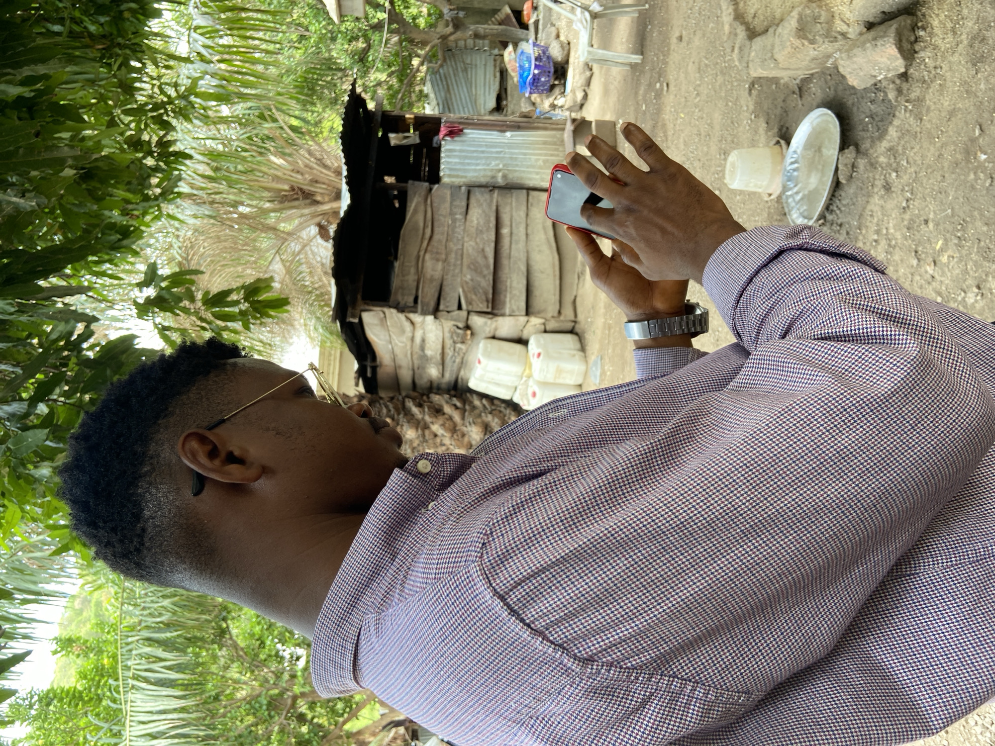 A man with dark hair and wearing glasses is seen from the side, holding a red smartphone, likely taking a photograph. He is wearing a button-down shirt with a small checkered pattern. The scene behind him appears to be a village or rural area. A partially built structure of wood and corrugated metal sheeting is visible. Several white plastic jerrycans are stacked nearby. A white plastic cup and metal plate are on the ground to the right. The foliage of trees is visible on the left side of the frame. The environment is consistent with a less developed area, and based on the prompt, this scene may be located in Mararaba, Nigeria.