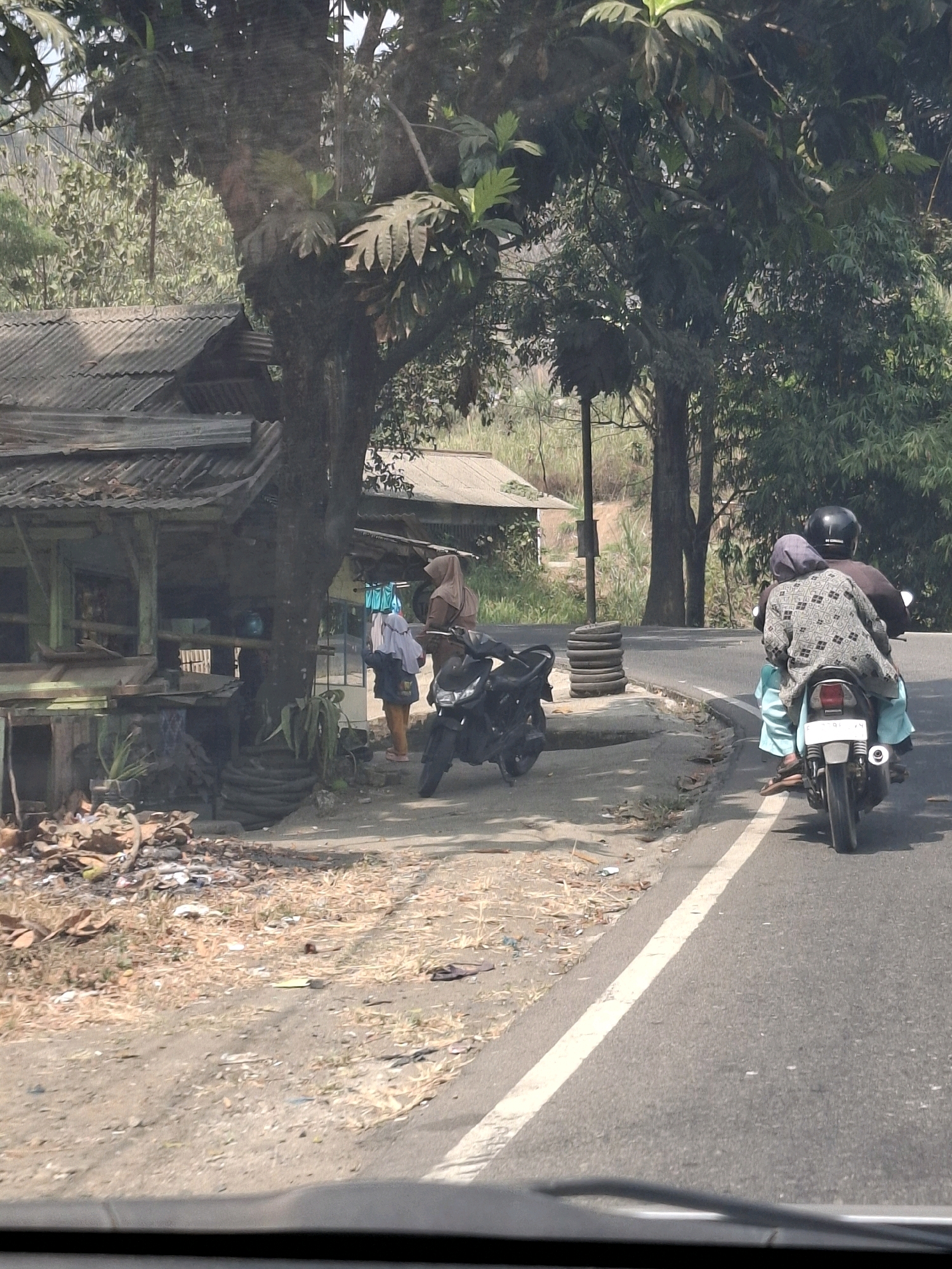 In Cijangkar, Indonesia, a roadside scene unfolds near a rudimentary structure. A person, wearing a headscarf and light-colored pants, stands next to a black motorcycle. Further down the road, two individuals are riding on another black motorcycle. The person in front wears a helmet and a dark top, while the passenger behind them is adorned in a patterned top and light blue pants, also with a headscarf. The environment consists of a paved road with a white line marking its edge, bordered by a dusty embankment littered with debris, dry vegetation, and discarded items. A weathered building with a corrugated metal roof is situated to the left, partially obscured by a large tree with dense foliage. In the background, more trees and vegetation suggest a rural or semi-rural setting. The lighting indicates it is daytime, likely with bright sunlight.