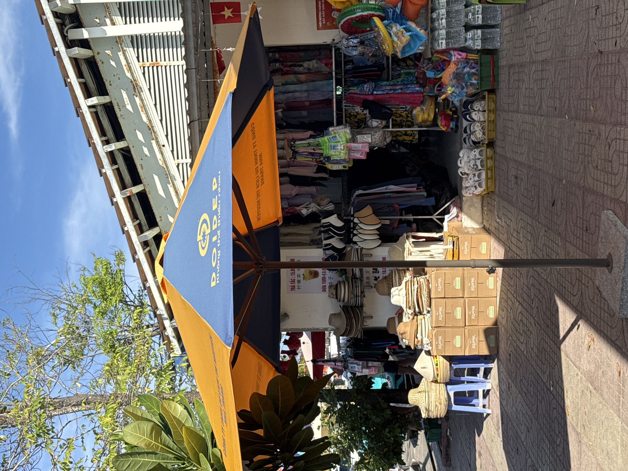 The image shows a store or market stall on a city street, likely located in Nam Nha Trang Ward, Vietnam, given the Vietnamese flag visible. The stall displays a variety of goods, including clothing, hats, shoes, and packaged items. A large blue and yellow umbrella stands in front of the stall, providing shade. The store appears to have a corrugated metal roof. There are stacked boxes of Aquafina water, small plastic stools, and some leafy plants near the bottom of the frame. The sidewalk is made of patterned tiles.