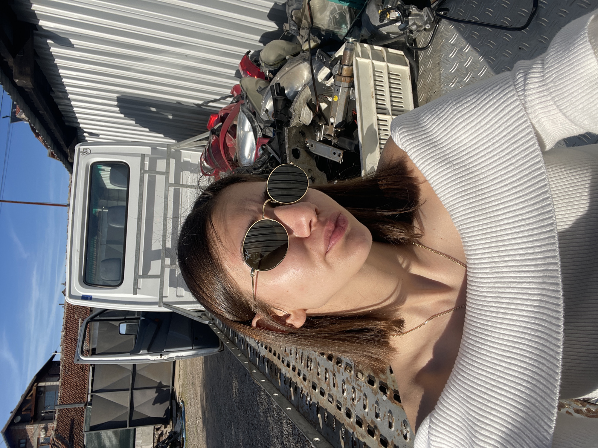 A young woman with straight brown hair, wearing round gold-framed sunglasses (with a visible Ray-Ban logo on one lens) and a white ribbed top, is captured in a bright, sunny outdoor setting in Sofia, Bulgaria. She is positioned in profile, her head slightly tilted, with a subtle pouting expression. A thin gold chain necklace is visible around her neck.
The background suggests an industrial or salvage yard. Immediately behind her is a dense stack of various vehicle components, including red tail lights, headlights, bumpers, and a white plastic crate, reflecting the intense sunlight. To her left, a white truck or van is parked, with its driver's side door open, revealing part of its interior.
Above and behind the truck and vehicle parts, a corrugated metal structure, likely a roof or wall, is prominent, casting sharp shadows. Beyond the immediate foreground, a clear blue sky extends, with distant glimpses of buildings featuring traditional tiled roofs, suggesting a surrounding suburban or rural landscape. The woman appears to be resting against or situated on a metal platform with a diamond plate texture and perforated holes, possibly a part of the truck bed or a similar structure within the yard. The strong, direct sunlight creates bright highlights and deep shadows throughout the scene, indicative of a clear, midday or early afternoon.