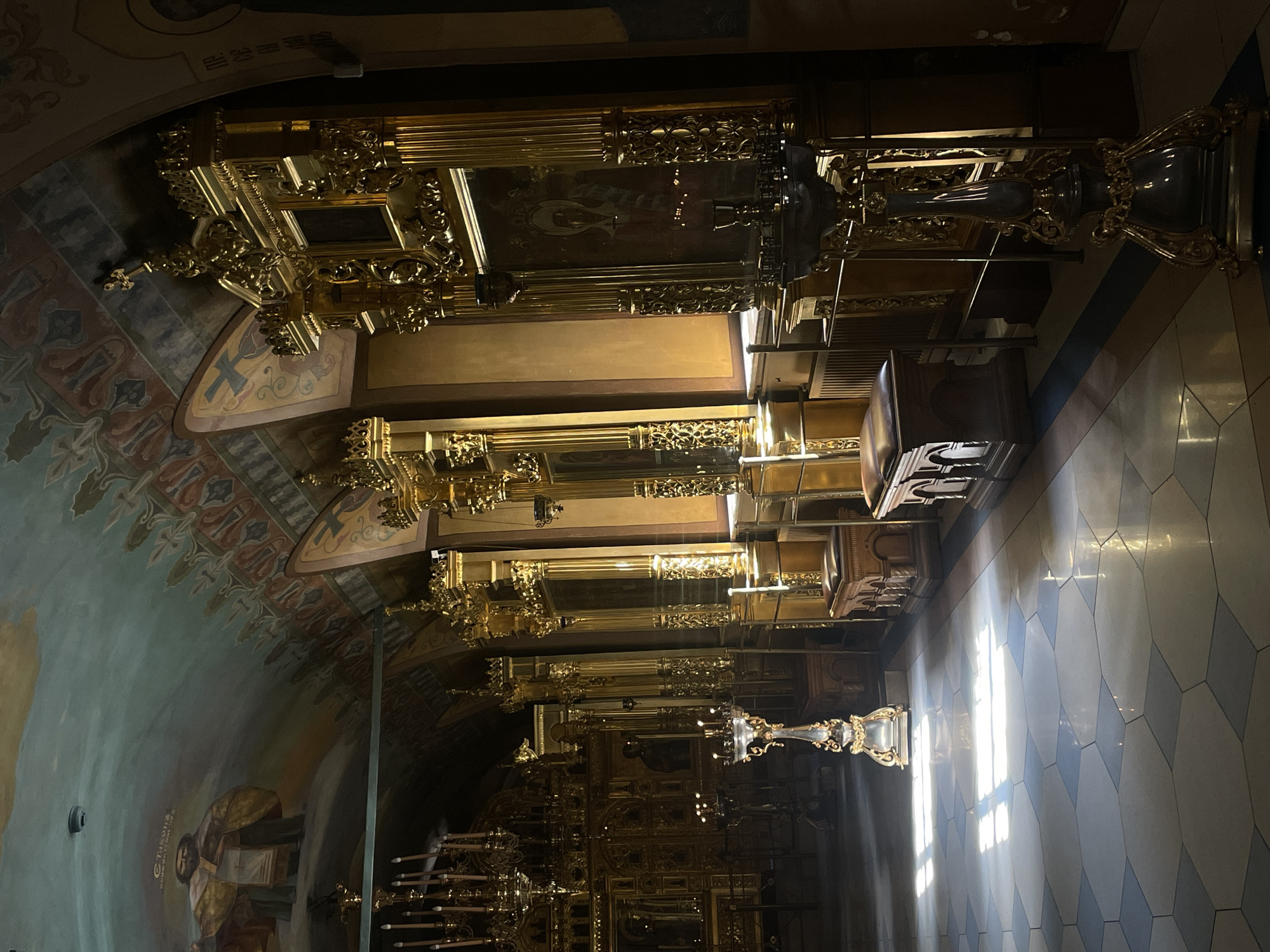 The image depicts the interior of a richly decorated Orthodox church, likely located in Moscow, Russia. The scene is dominated by a gilded iconostasis, a wall of icons and religious paintings separating the nave from the sanctuary. Ornate gold detailing adorns the structure, featuring intricate carvings, columns, and multiple tiers of religious imagery. Light streams in from a window on the right, illuminating the checkered floor tiles. The left wall displays a fresco or mural depicting religious figures and decorative patterns. In the foreground, there are candelabras holding multiple candles.