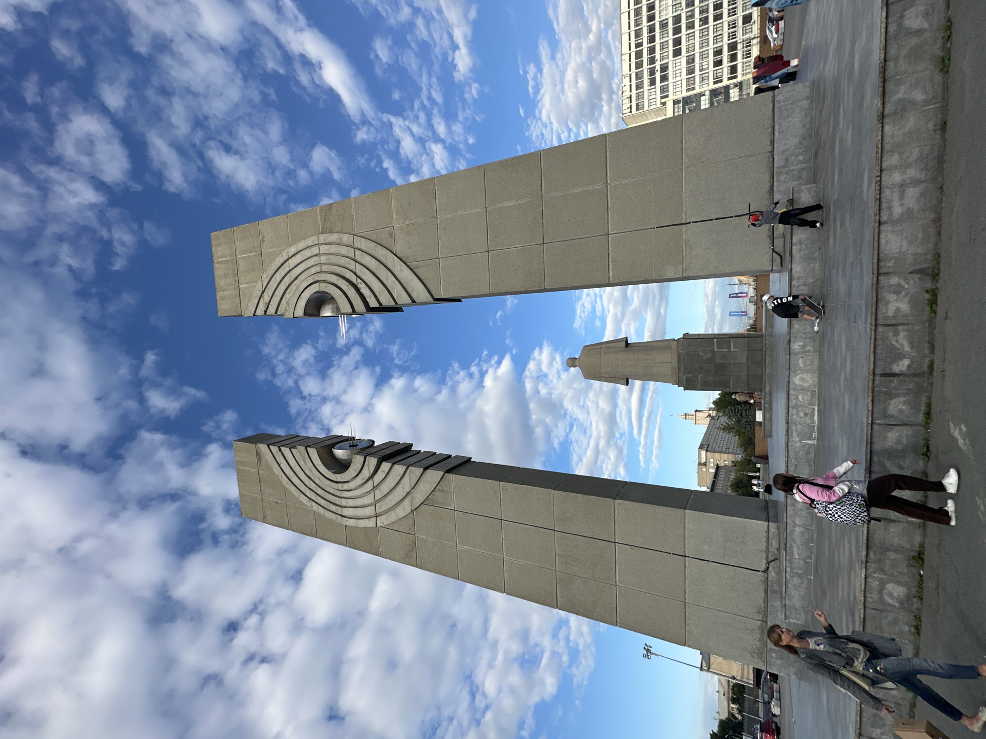 In an urban plaza under a partly cloudy sky, two imposing, sloped concrete pillars dominate the foreground. The pillars are textured with a series of concentric, raised arcs that converge towards the top of each structure. A tall, solid statue of a figure standing with its back to the viewer is positioned between and behind the pillars.
In front of the pillars, several individuals are present. To the left, a woman with long brown hair walks away from the camera, wearing a grey jacket, a t-shirt, and blue jeans, carrying a beige bag and a cardboard box. Next to her, a young person with dark hair and a pink top walks with a large checkered backpack. Further to the right, a child wearing a white helmet and black shorts is on a skateboard, while another child in a red hat and black pants stands with a stick.
In the background, a large, multi-story white building with many windows is visible, along with other city buildings and some trees. Cars are parked on a street to the far left and right of the plaza. The ground is paved with large gray tiles, and a low concrete barrier runs along the edge of the plaza.