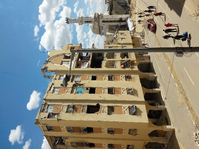 The image depicts a damaged urban street scene in Khan Yunis, Palestinian Territory, under a bright blue sky with scattered white clouds. In the foreground on the left, a multi-story, light-colored residential building exhibits significant structural damage, including numerous impact holes, shattered windows, and partially collapsed sections on its upper floors and roofline. Some balconies feature hanging laundry and debris. The ground floor of this building is supported by arched columns, beneath which two vehicles are partially visible.
A paved road with a central white double solid line runs through the scene. Along the right side of the road, approximately 15-20 individuals, mostly adults, walk away from the viewer, some carrying bags or bundles. A red sedan is parked or stopped on the right shoulder of the road. In the mid-foreground, a tall grey utility pole stands centrally, partially obscuring the street.
In the background on the right, a mosque with a tall white minaret and a dome is visible, also appearing damaged with rubble at its base and surrounding structures. The general environment indicates extensive destruction, with debris scattered along the sidewalks and edges of the road.