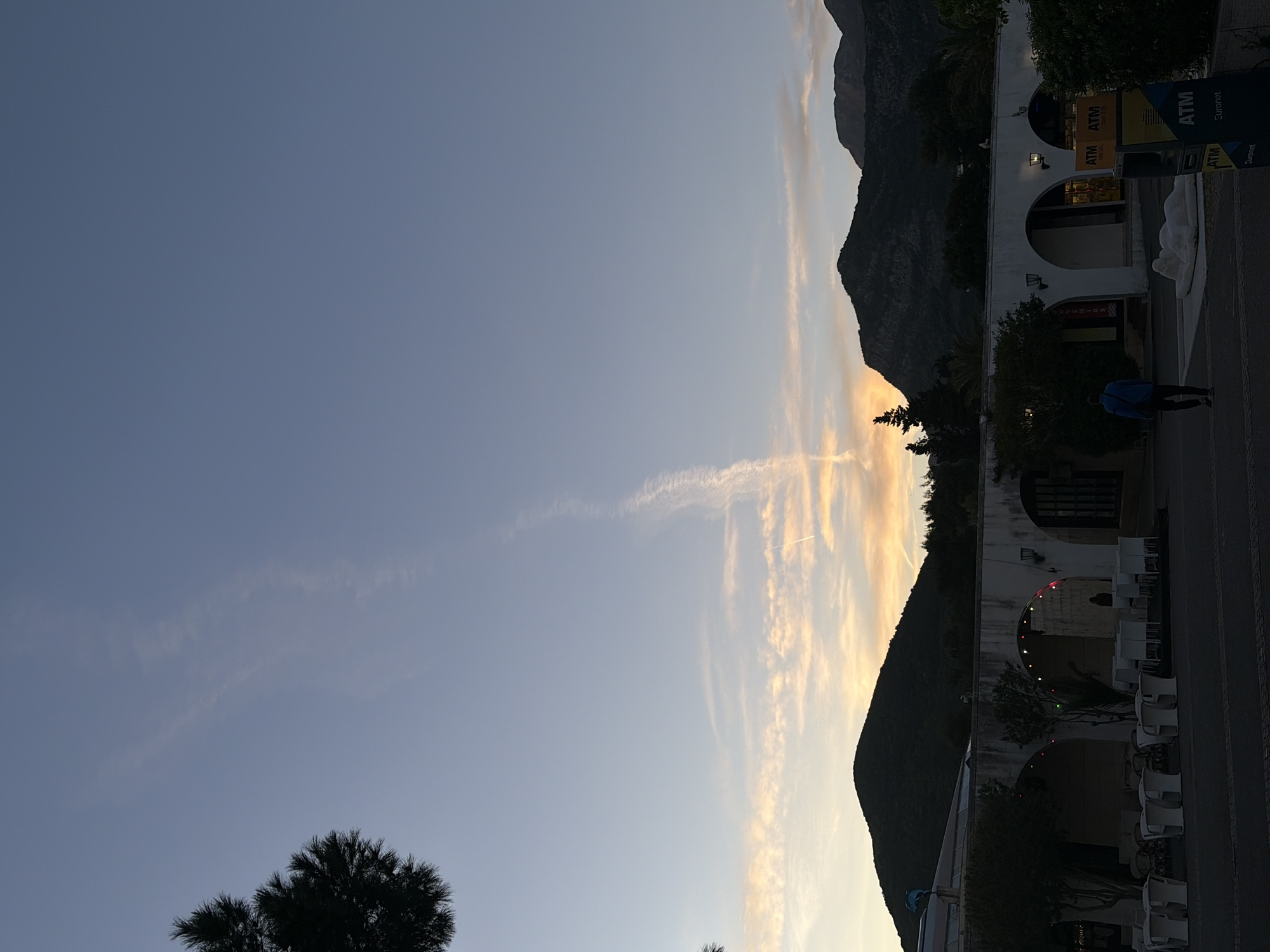 The image captures an outdoor scene at dusk or dawn in Budva, Montenegro, characterized by a serene sky transitioning from blue-grey on the left to warm golden-orange hues on the right horizon. Wispy clouds and faint contrails stretch across the upper sky. A dark, mountainous landscape forms the backdrop, largely silhouetted against the bright horizon. In the mid-ground, a series of white, arch-fronted buildings are visible, also appearing as dark forms against the sky's glow.
In the foreground, a paved street or walkway extends across the lower part of the frame. On the far left, a dark tree is silhouetted against the sky. Further right on the street, a person wearing a blue shirt and dark pants walks away from the viewer. Prominently placed near the street are two yellow and black ATM signs, clearly displaying the text "ATM", "Euronet", and "24h". The buildings feature multiple arched entrances, with some interior lights and decorative string lights (red, green, blue) visible under one arch. Several white outdoor chairs and tables are arranged along the street, suggesting a dining or relaxation area. The overall atmosphere is tranquil, with the natural light of the sky contrasting with the shadowed terrestrial elements.