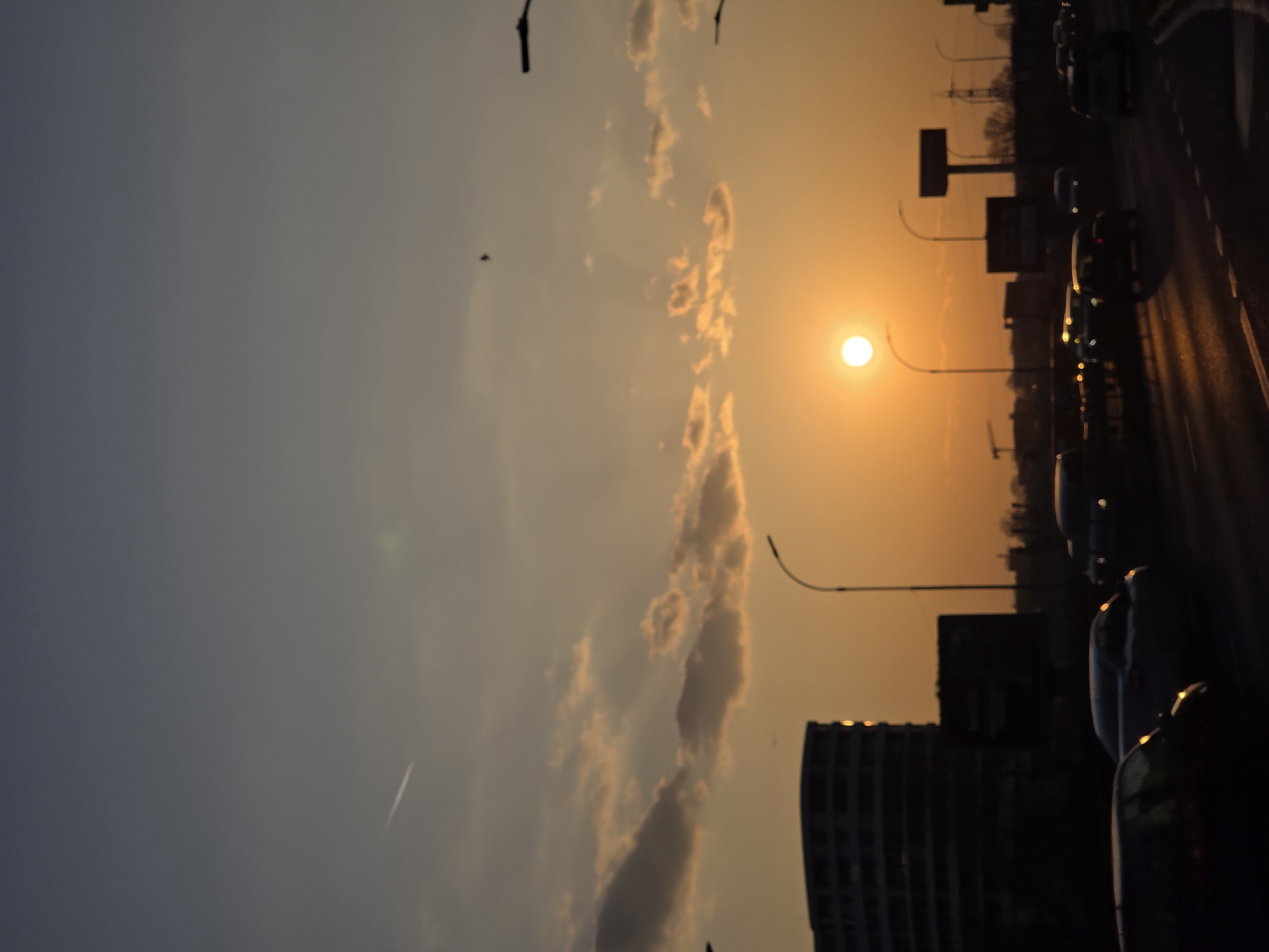 The image presents an urban or suburban landscape within Varna, Bulgaria, observed at sunset. The sun, a distinct bright orange-yellow sphere, is positioned low in the central-right portion of the sky, radiating an amber light that illuminates the lower atmosphere. The sky transitions from a dark blue-grey at the top to a vibrant orange-yellow nearer the horizon, containing scattered dark and golden-edged cumulus clouds, with some exhibiting visible crepuscular rays. A thin, white aircraft contrail streaks across the upper-left quadrant, and a small, dark bird is captured in flight in the upper-mid-right.
In the foreground and midground, a multi-lane asphalt road with visible white lane markings extends diagonally from the lower-left towards the right. Numerous passenger vehicles are present, mostly appearing as dark silhouettes with illuminated headlights and taillights, reflecting the sunset's glow and indicating active traffic flow.
The background is dominated by a silhouetted urban skyline. A prominent dark, multi-story building with a rounded architectural facade is visible on the left, displaying several points of sunlight reflection from its windows. To its right, various other dark building structures, an apparent construction crane, and power line pylons are silhouetted against the bright horizon. Multiple streetlights with curved arms are positioned along the roadway and skyline. A large, T-shaped billboard structure is also visible in silhouette on the right side of the road, beyond the immediate traffic.