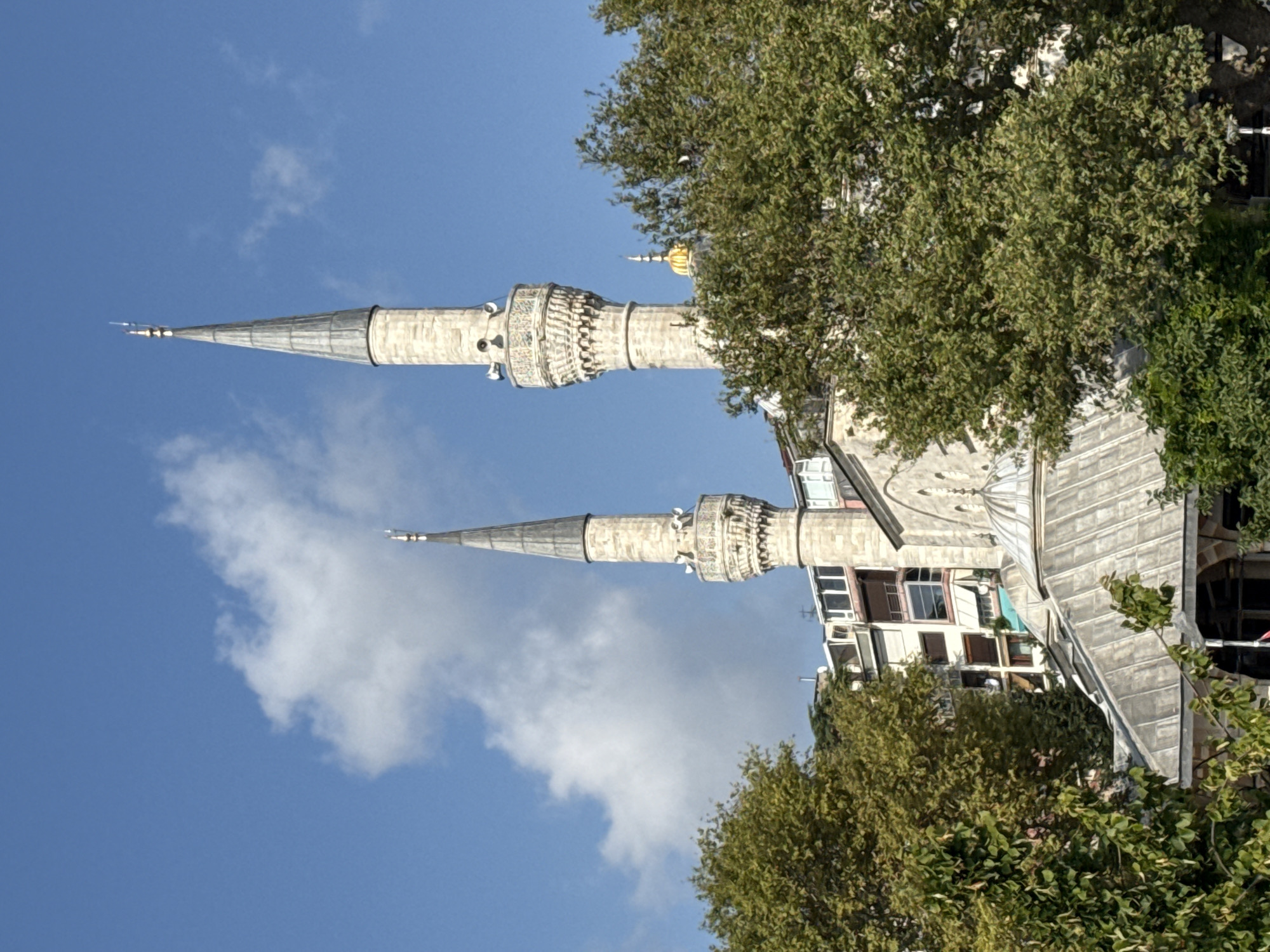 The image shows two minarets of a mosque in Üsküdar, Turkey, against a blue sky with scattered white clouds. The minarets are tall, slender, and made of stone. They have balconies and are topped with conical spires. In front of the minarets, there is a domed roof structure, also made of stone. Trees with green foliage partially obscure the view of the mosque and the surrounding buildings, which appear to be apartments. The scene is lit by sunlight, casting shadows and highlighting the architectural details.