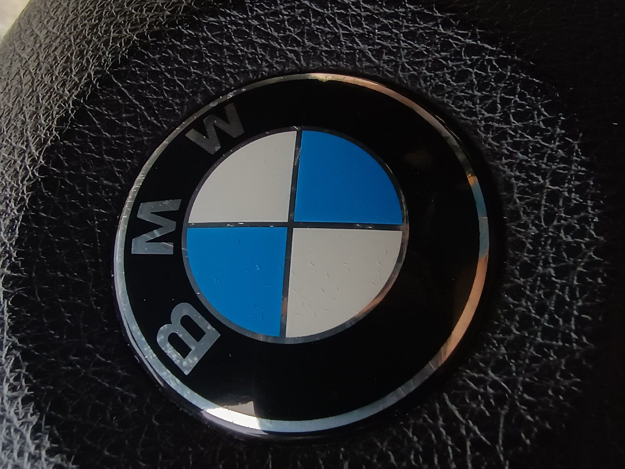 The image shows the BMW logo affixed to a textured black surface, likely leather or a leather-like material, suggesting it is part of a car interior, possibly the steering wheel or dashboard. The logo is circular, featuring a black outer ring with the letters "BMW" in silver/gray. The central circle is divided into four quadrants, alternating between blue and white. No people are visible in the image. The location of the image within the city of Plovdiv, Bulgaria, cannot be determined from the given image.