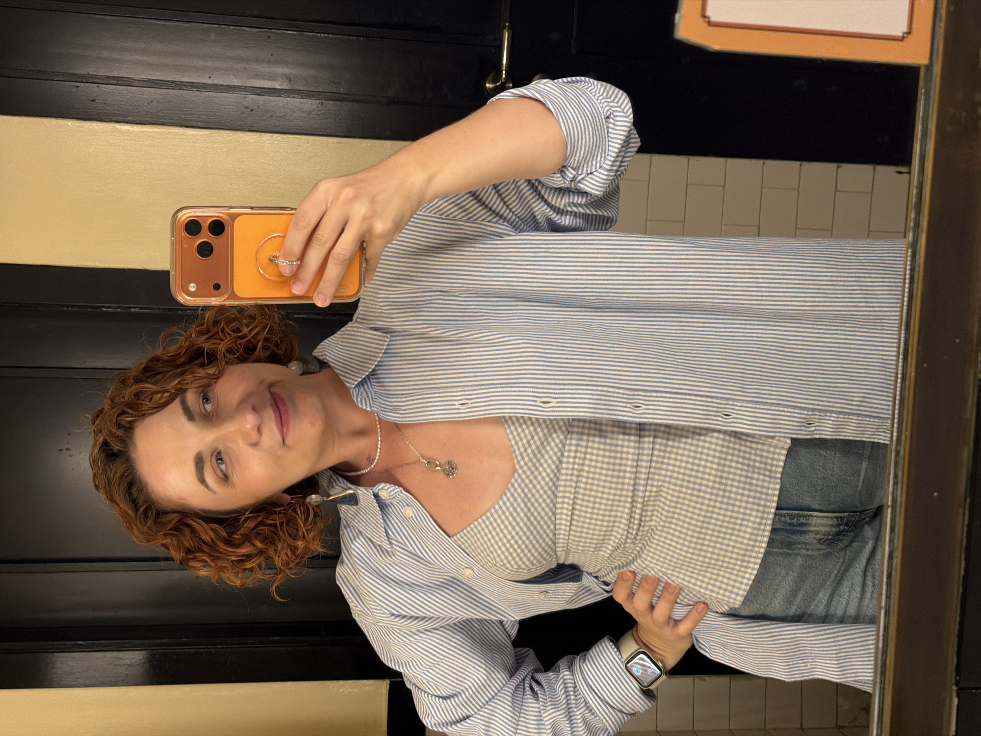 A woman with red curly hair and blue eyes is taking a selfie in a bathroom. She is holding an orange phone with a ring stand on the back. She is wearing a blue and white striped shirt over a light blue and white gingham top, with blue jeans and a silver necklace with a circular pendant. She has a watch on her left wrist. The bathroom has horizontal stripes of black and tan paint. There is a sign above her head in the mirror's reflection. The bathroom has subway tile. There is no indication the photo was taken in Tel-Aviv, Israel.