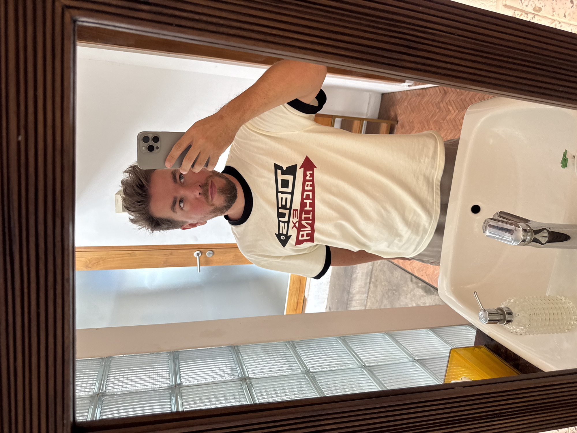 A man is taking a selfie in a bathroom mirror. He is wearing a beige t-shirt with black trim and the word "DEUS EX MACHINA" printed in red and black. The bathroom has a white sink, brick flooring, and a window with glass blocks. The photo was likely taken indoors. Based on the information given that location is in the Ubud District, Indonesia.