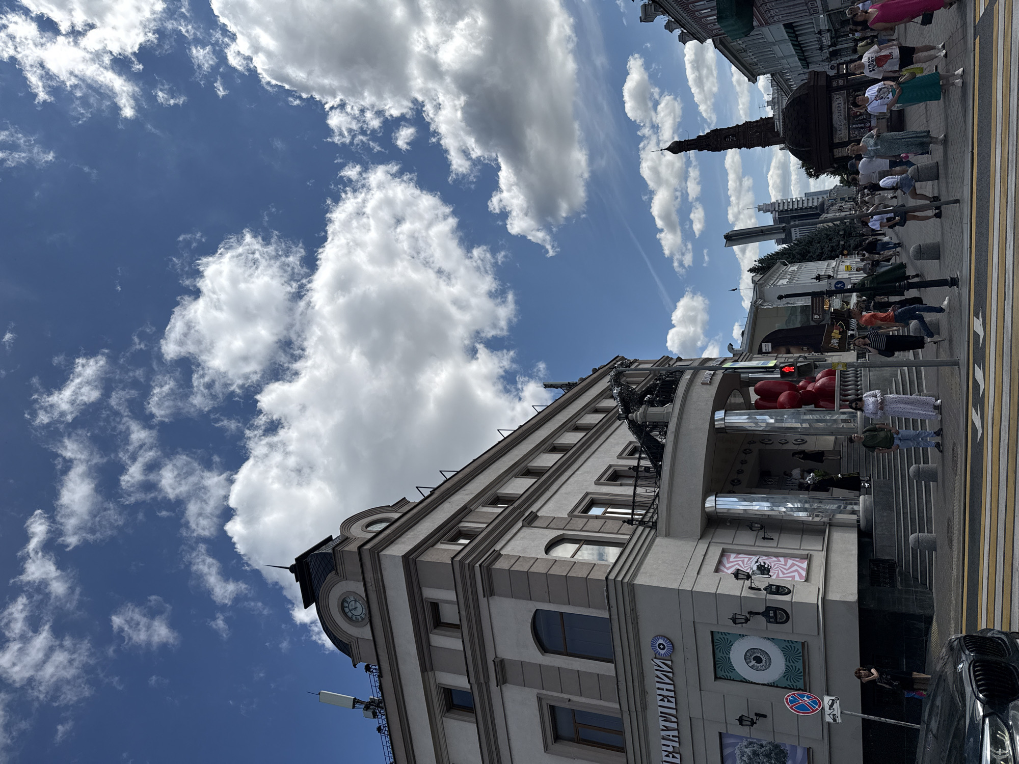 The image shows a view of Bauman Street in Kazan, Russia, on a sunny day with scattered clouds. The street is paved with cobblestones and lined with historic buildings. In the foreground, a black car is parked on the left side, and a pedestrian crossing with yellow stripes is visible. Several people are walking on the sidewalks, dressed in casual summer clothing.
On the left, a grand building with a clock tower dominates the scene. It features classical architecture with columns, arched windows, and decorative moldings. A sign on the building reads "Впечатлений," which translates to "Impressions." On the right side of the street, a tall, slender tower with a dark, ornate structure at its base stands out against the sky. Further down the street, modern buildings with glass facades are interspersed with older structures. A red traffic light is visible in the middle of the street. The overall atmosphere is that of a lively urban environment on a pleasant day.