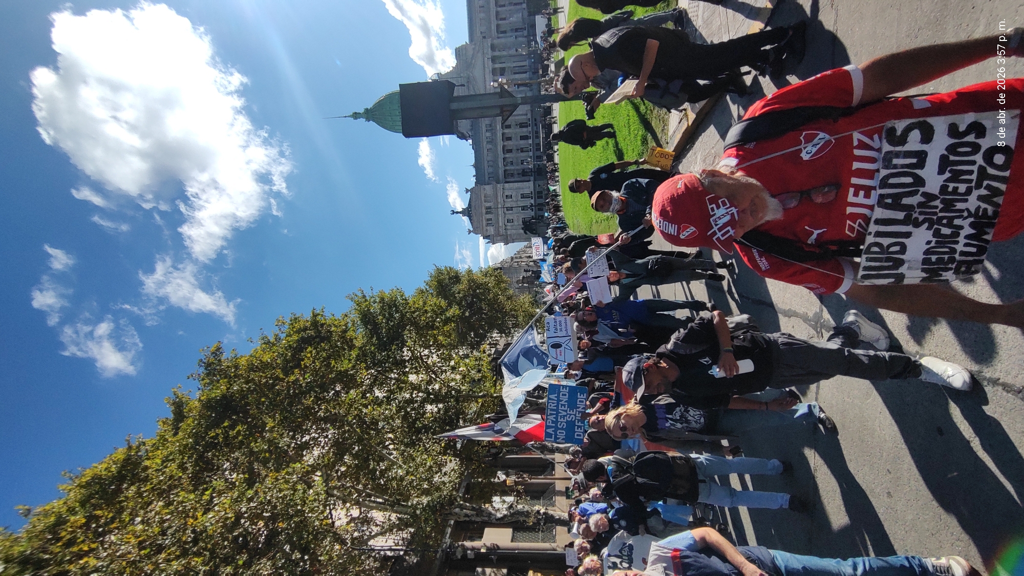 The image shows a daytime protest in Buenos Aires, Argentina. A crowd of people is gathered on a paved area, likely a street or plaza. Many are carrying signs and banners with messages written in Spanish, including "LA PATRIA.... NO SEVENDE SE DEFIENDE" and "JUBILADOS SIN MEDICAMENTOS." Some individuals are lying on the ground. One person is wearing a red jersey and cap with logos, and a sign attached to their back. In the background, a large building with a dome and classical architecture is visible. Trees line the edge of the gathering. The sky is blue with scattered clouds. The timestamp in the top right corner reads "8 de abr. de 2026 3:57 p. m."
